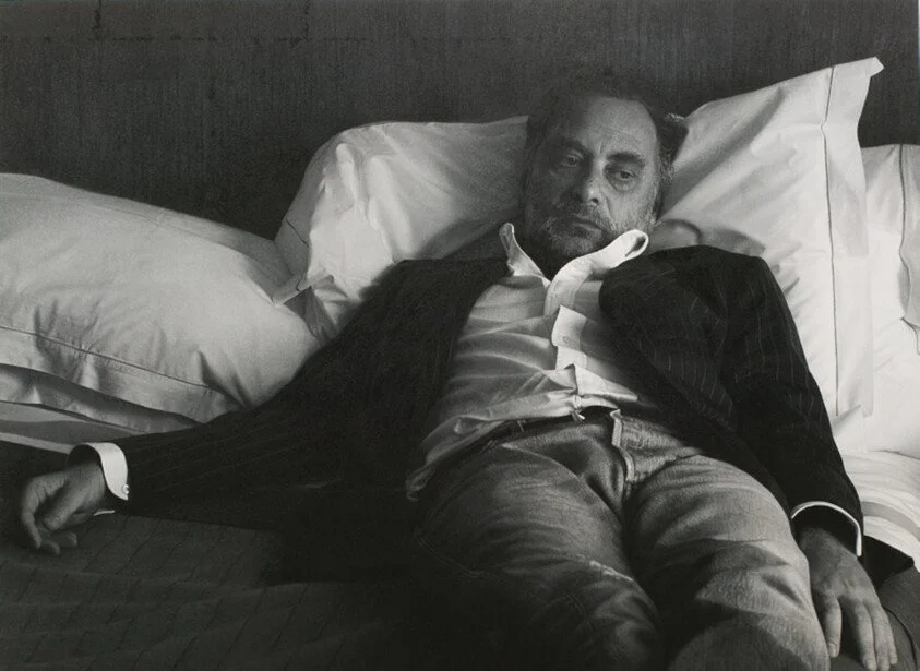 A man lying on a bed with pillows, wearing a suit jacket, shirt, and jeans, appearing to be asleep or resting.