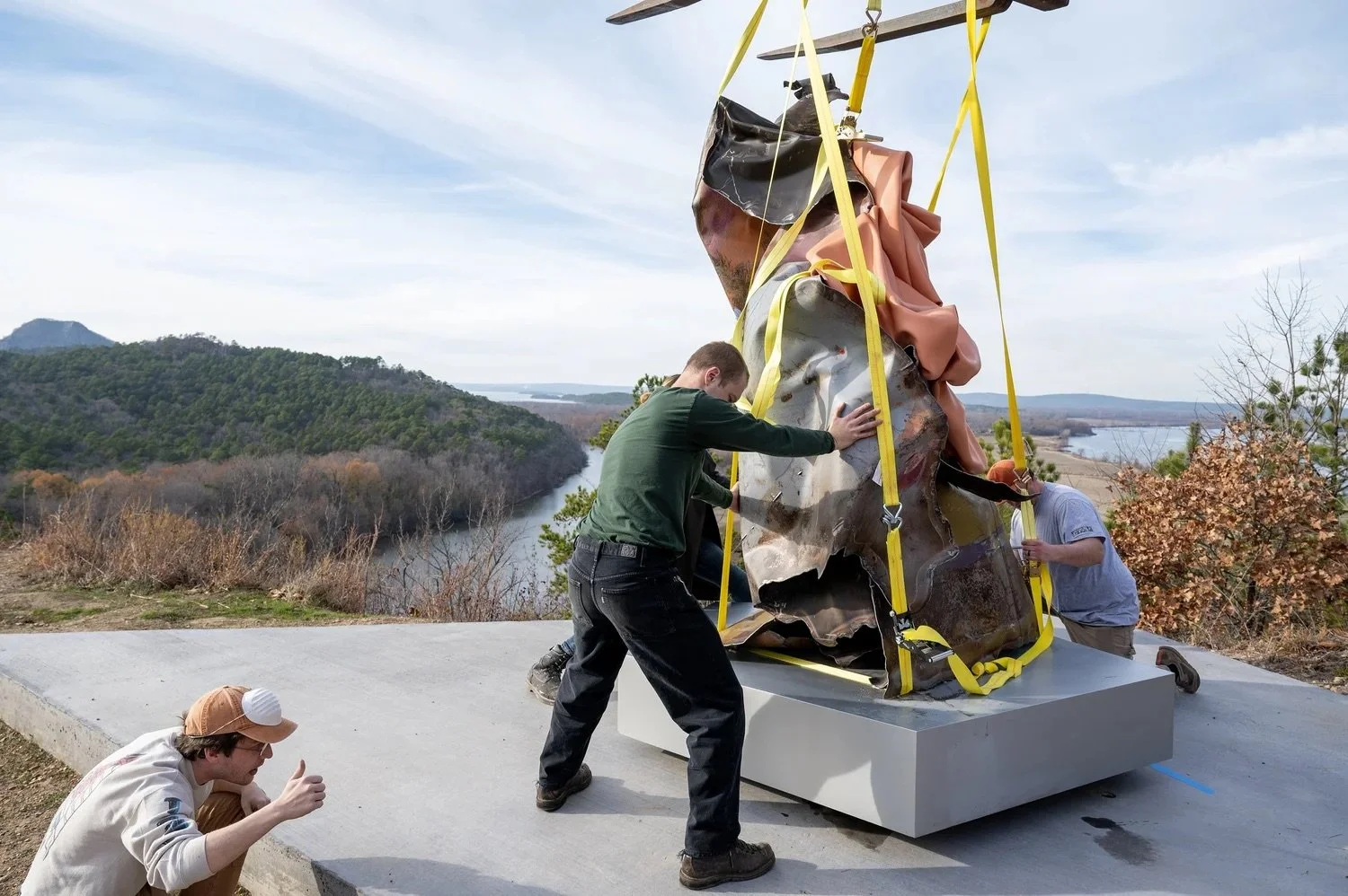 A group of people installing a large, abstract sculpture outdoors on a concrete platform overlooking a river and wooded landscape.
