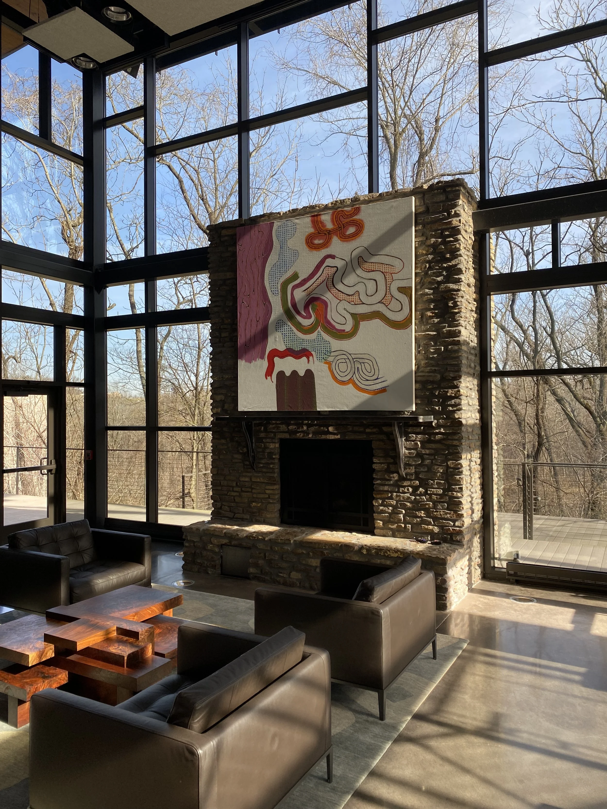 Interior of a modern building with large glass windows, a stone fireplace, and contemporary furniture, including black leather sofas and a wooden coffee table, with leafless trees visible outside.