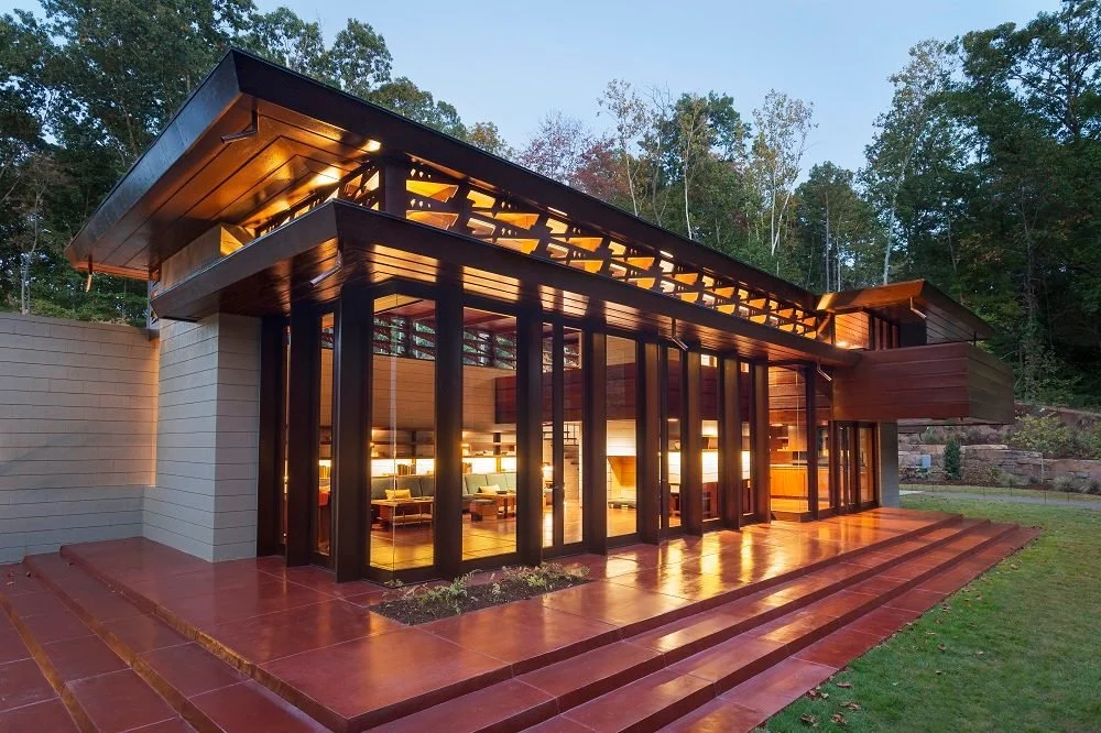 Modern house with large glass windows, wooden exterior, and a tiled patio in a wooded area, illuminated from inside during evening.