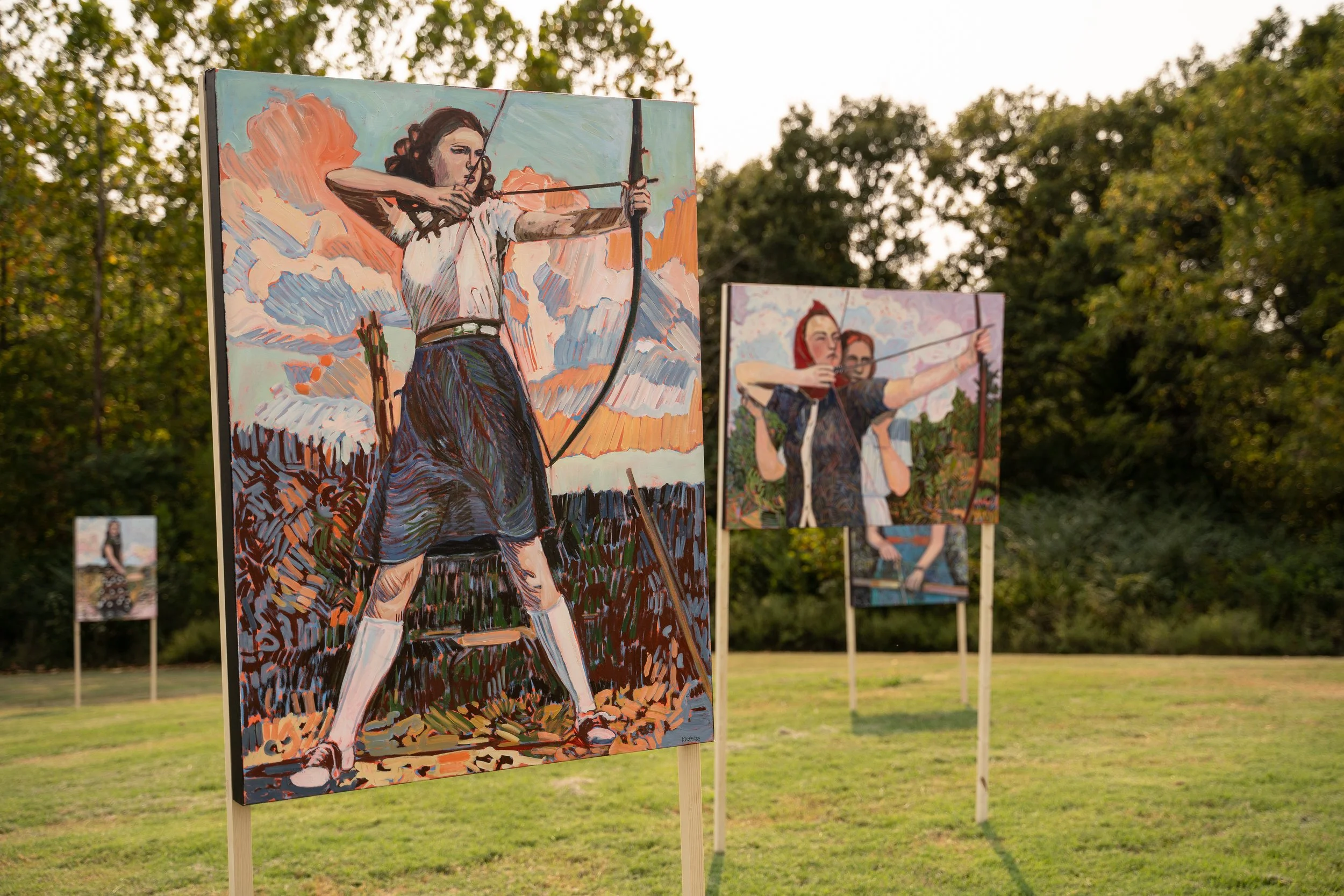 A painting of a woman with curly hair aiming a bow and arrow, with a sky filled with orange and light blue clouds background, displayed outdoors on an easel.
