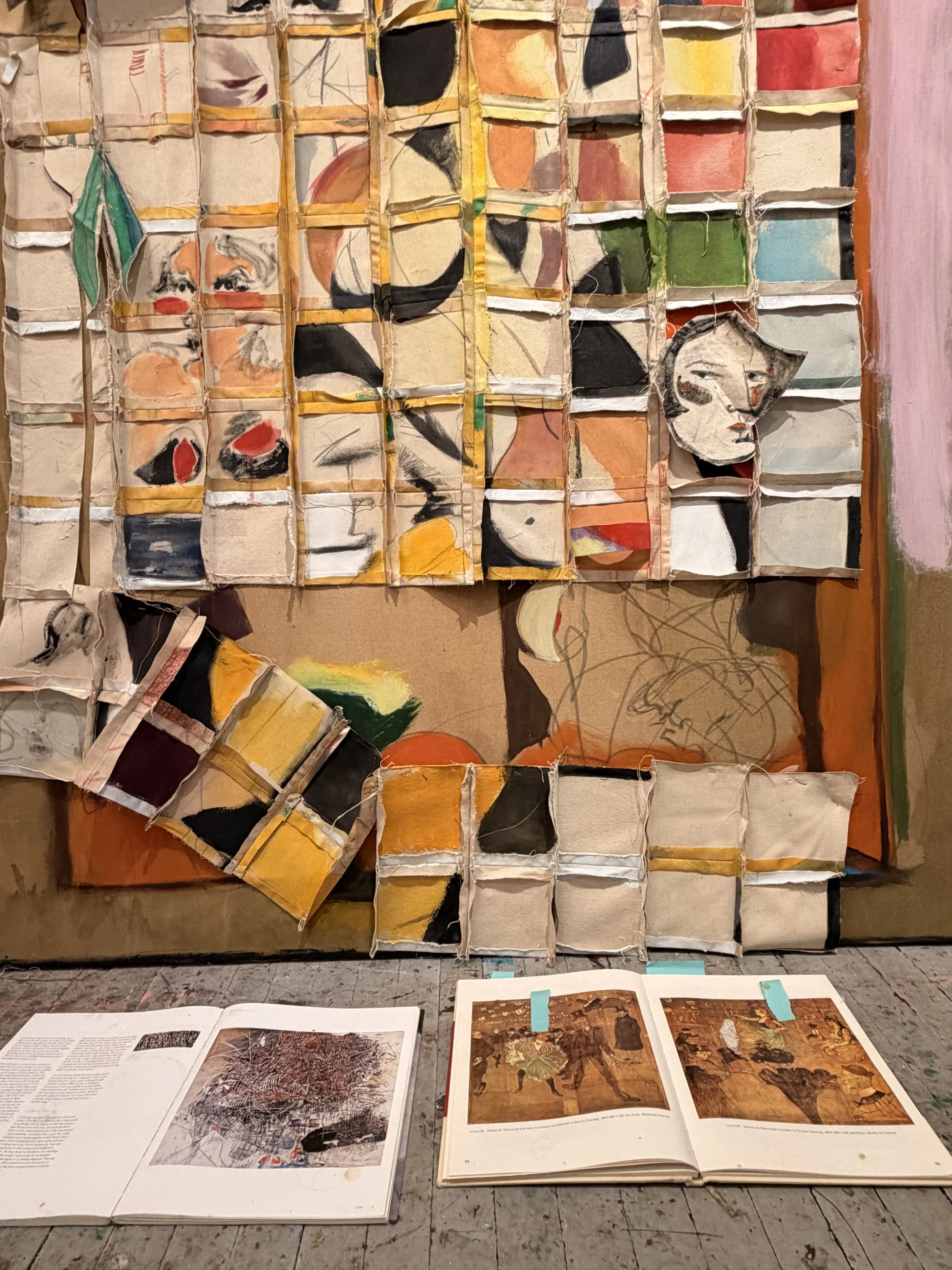 A mixed media art installation with a grid of fabric and paint pieces on the wall and open large art books with images of trees and artwork on the table in front.