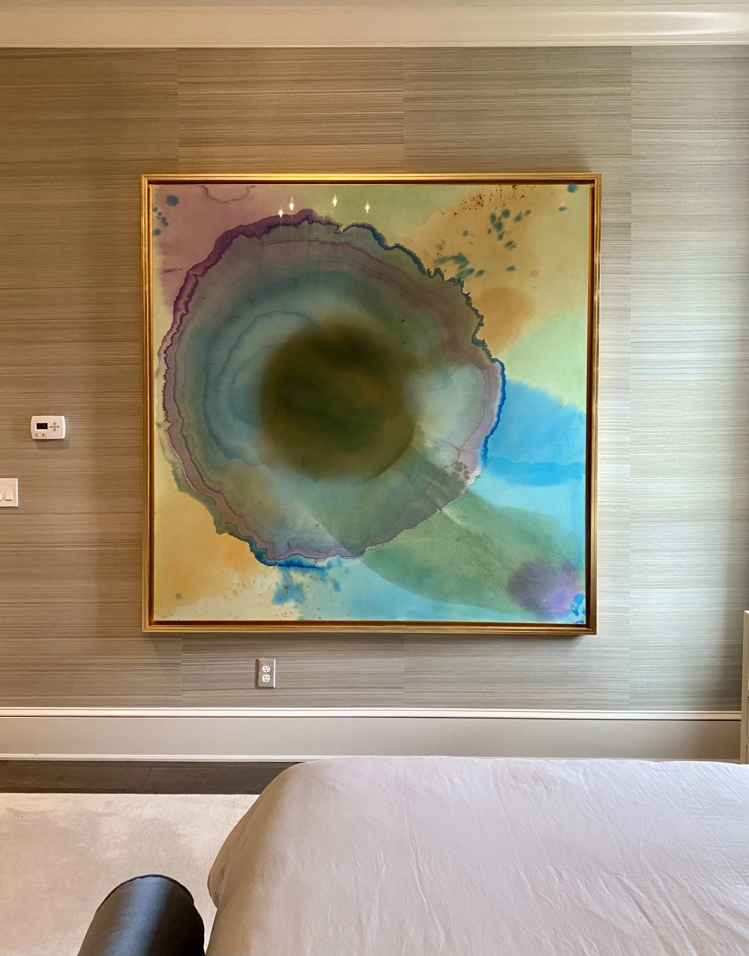 A large abstract watercolor painting with shades of purple, green, blue, and yellow on a beige wall in a room.