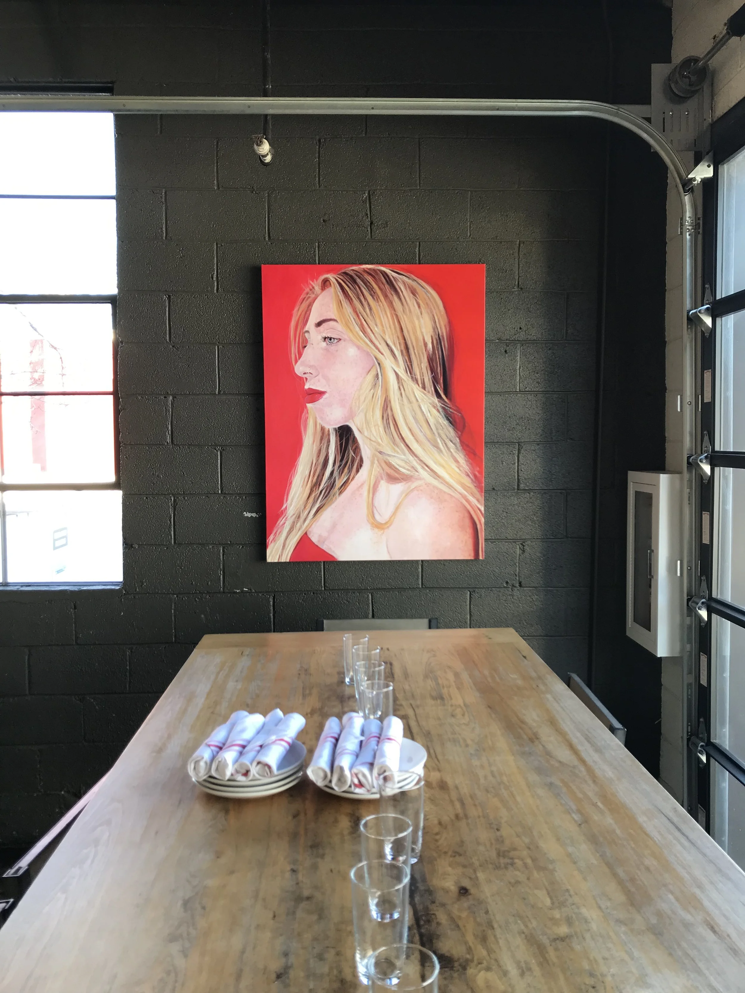 Interior of a modern restaurant or cafe with a long wooden table set with clear glasses and a plate of rolled white napkins. A colorful painting of a blonde woman on a red background hangs on a dark brick wall, with a window to the left allowing natural light to enter.