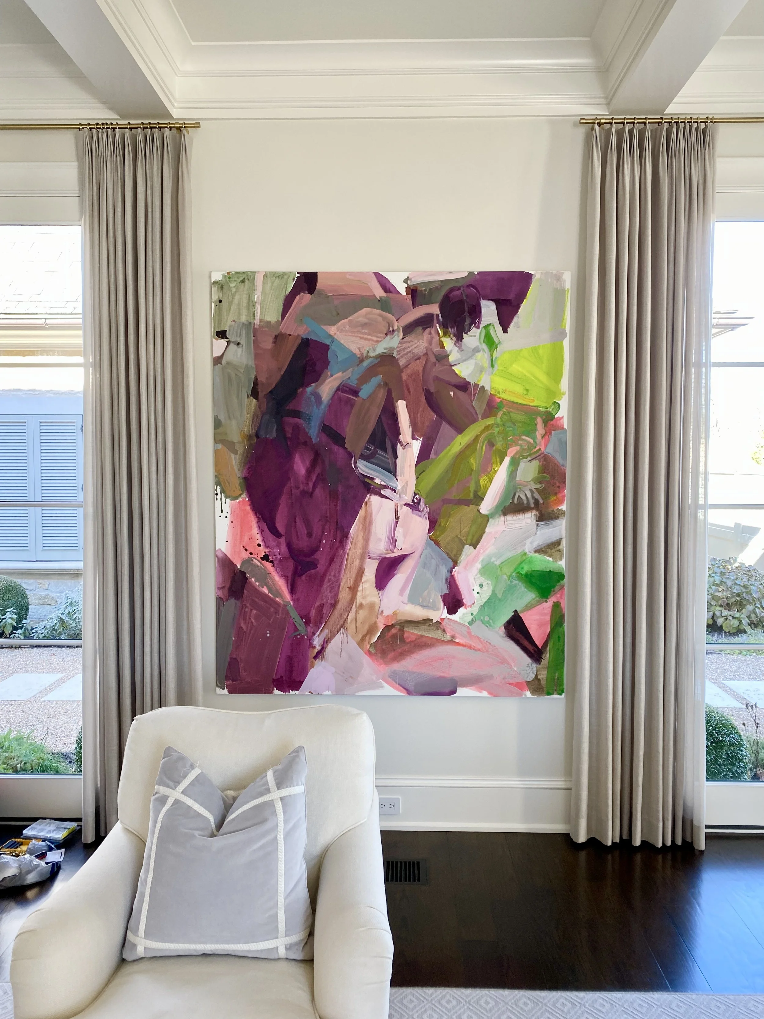 A colorful abstract painting hangs on a wall between two large windows with beige curtains. In front of the painting, there is a white armchair with a shiny silver pillow.