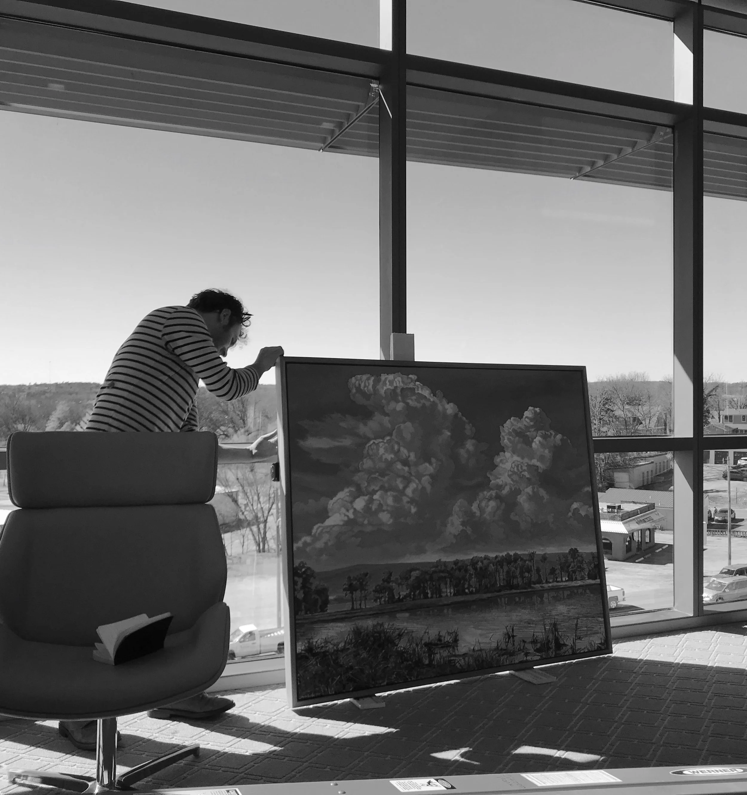 Black and white photo of a modern building with large windows. Two people are sitting at a table indoors, near a window. Outside the window, there are trees, a parking lot with cars, and a building in the distance.