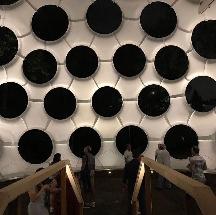 Interior design featuring a large wall with circular black panels arranged in a honeycomb pattern, with several people standing and conversing in front of the wall.