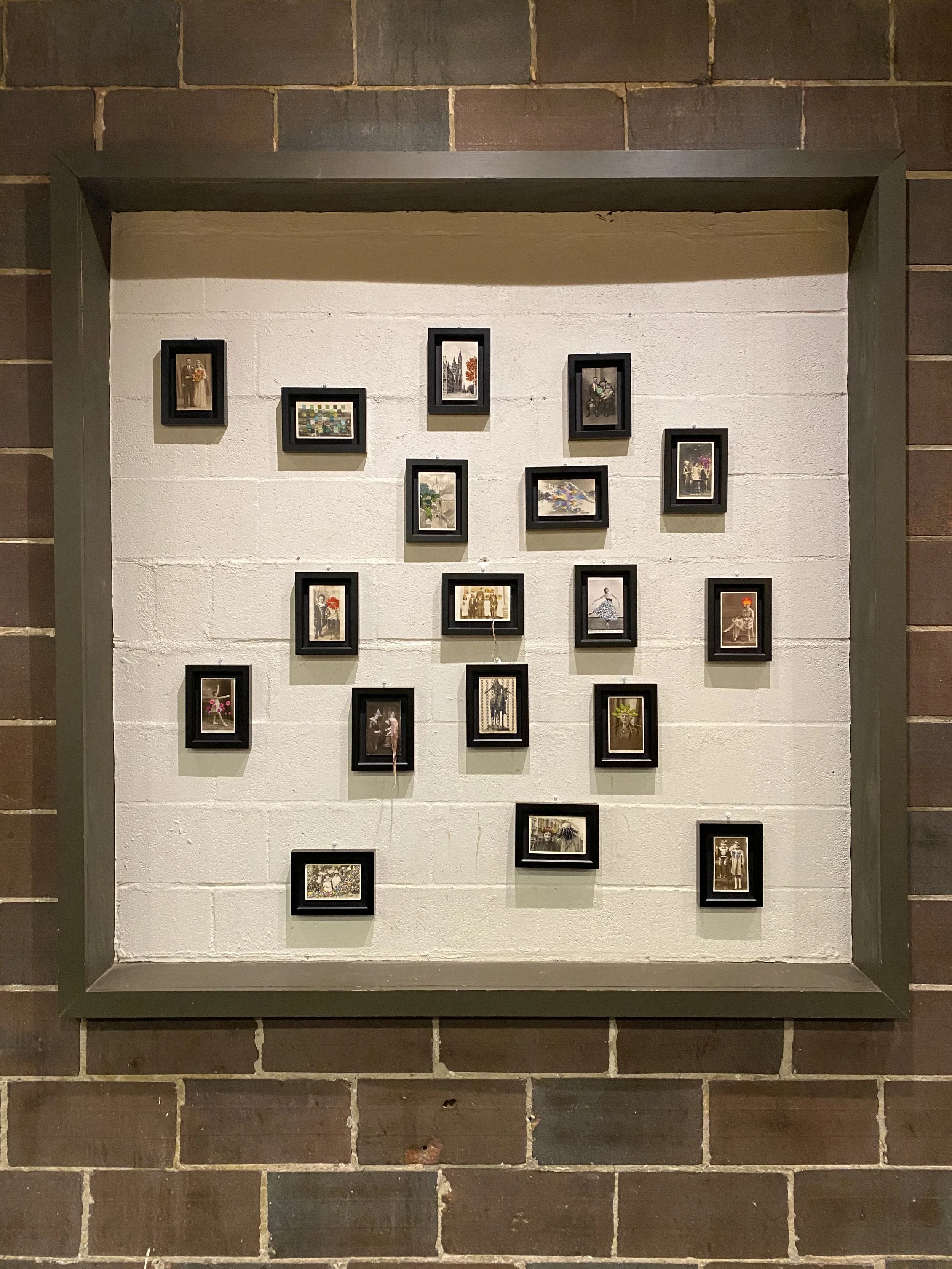 A wall display of framed photographs and artwork arranged in a grid pattern inside a black frame. The wall is painted light-colored, possibly white or cream, with a brick background surrounding the display.