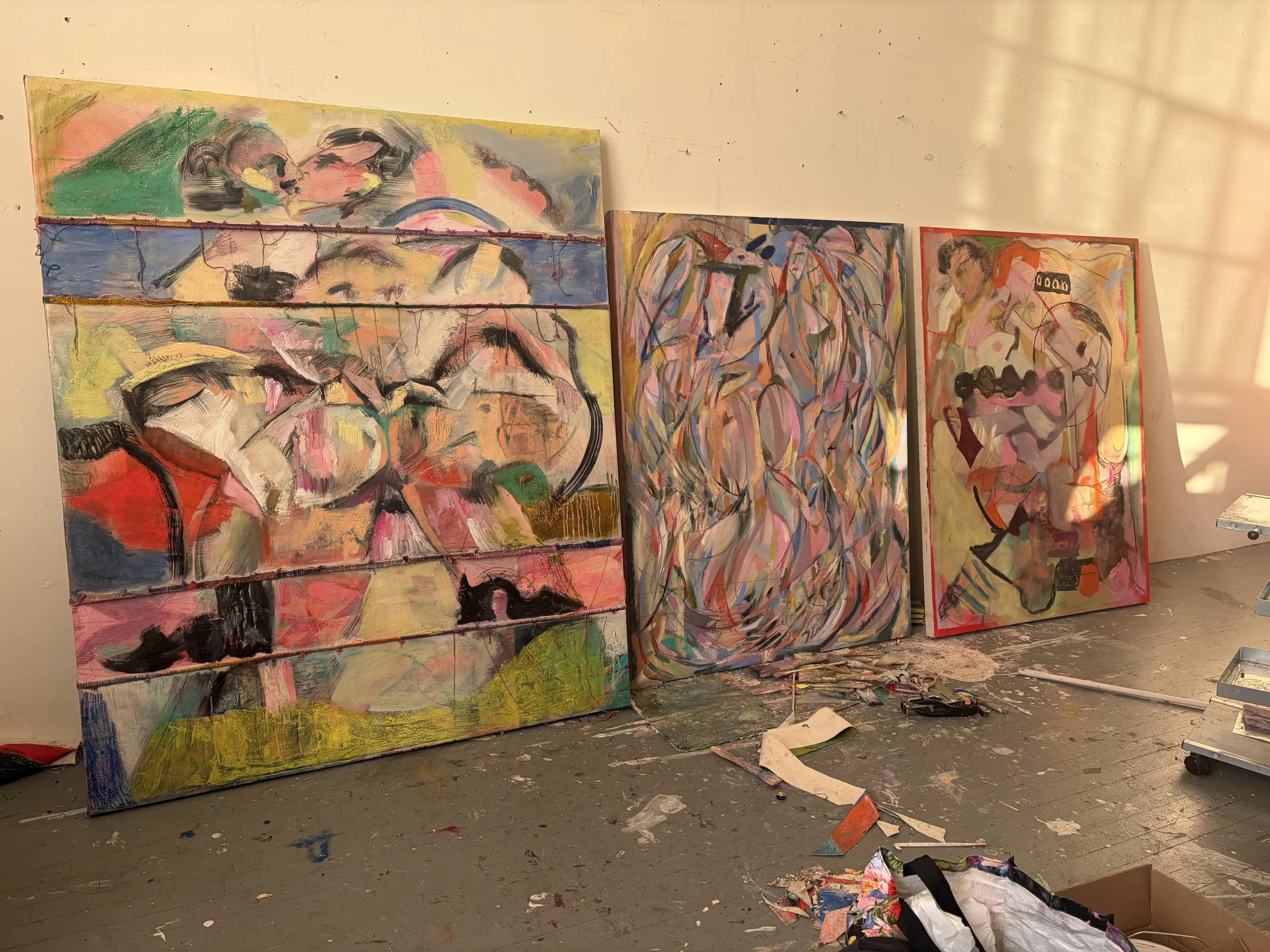 Multiple abstract paintings on a wall in an art studio with paint splatters on the floor and scattered painting supplies.