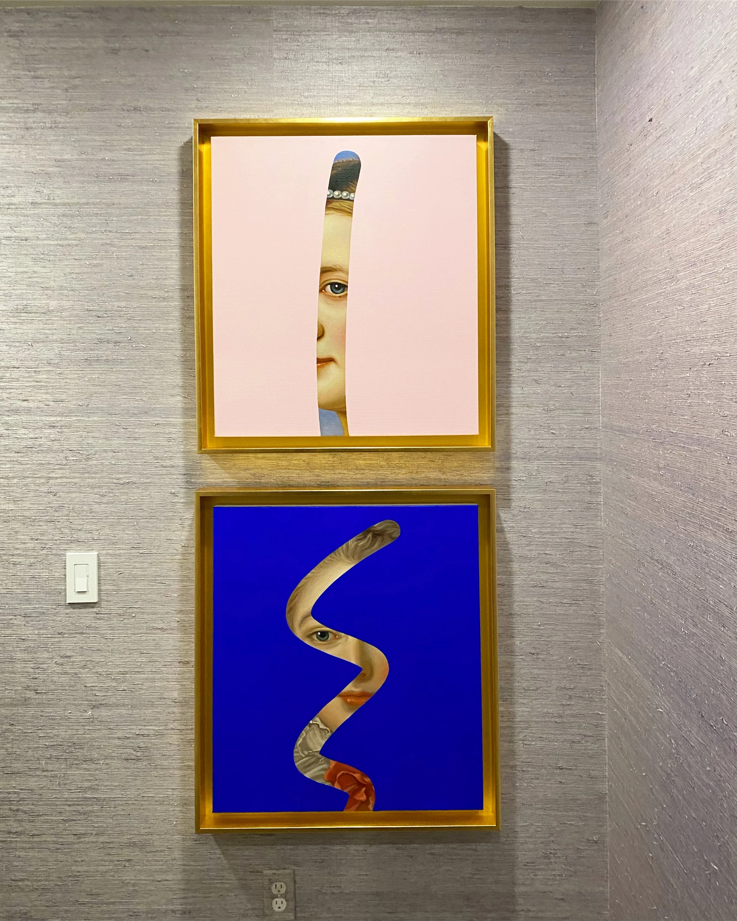 Two art pieces with human faces, one pink and one blue, featuring cutouts revealing portions of faces inside each, hanging on a textured grey wall.