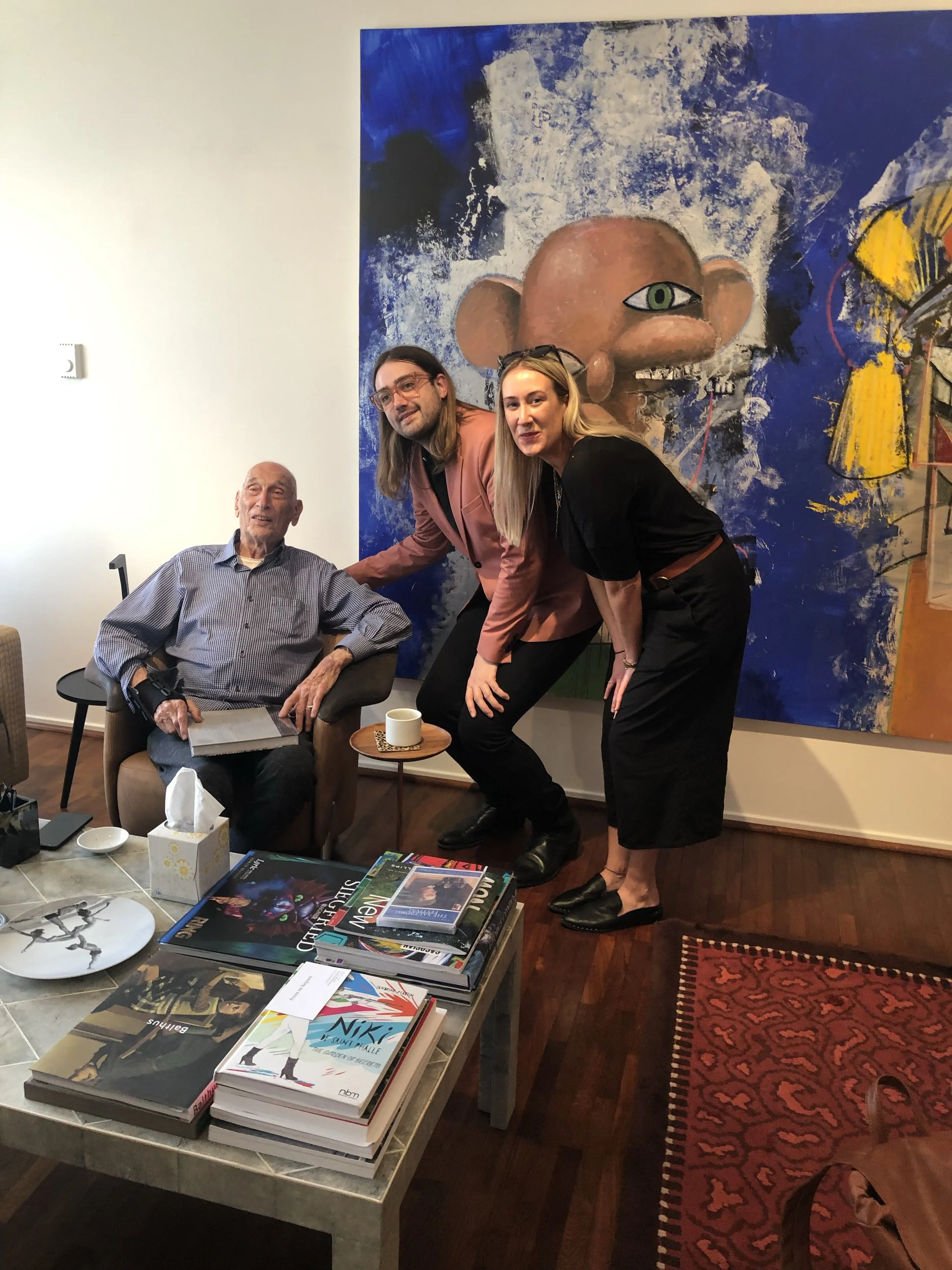 Three people are gathered in an art gallery with a large colorful mural of a surrealist face with a single eye behind them. The elderly man is seated in a chair, holding a book, with a table of books and magazines in front. Two women stand next to him, smiling and leaning slightly forward. The room has wooden floors, a patterned rug, and various books on the table.