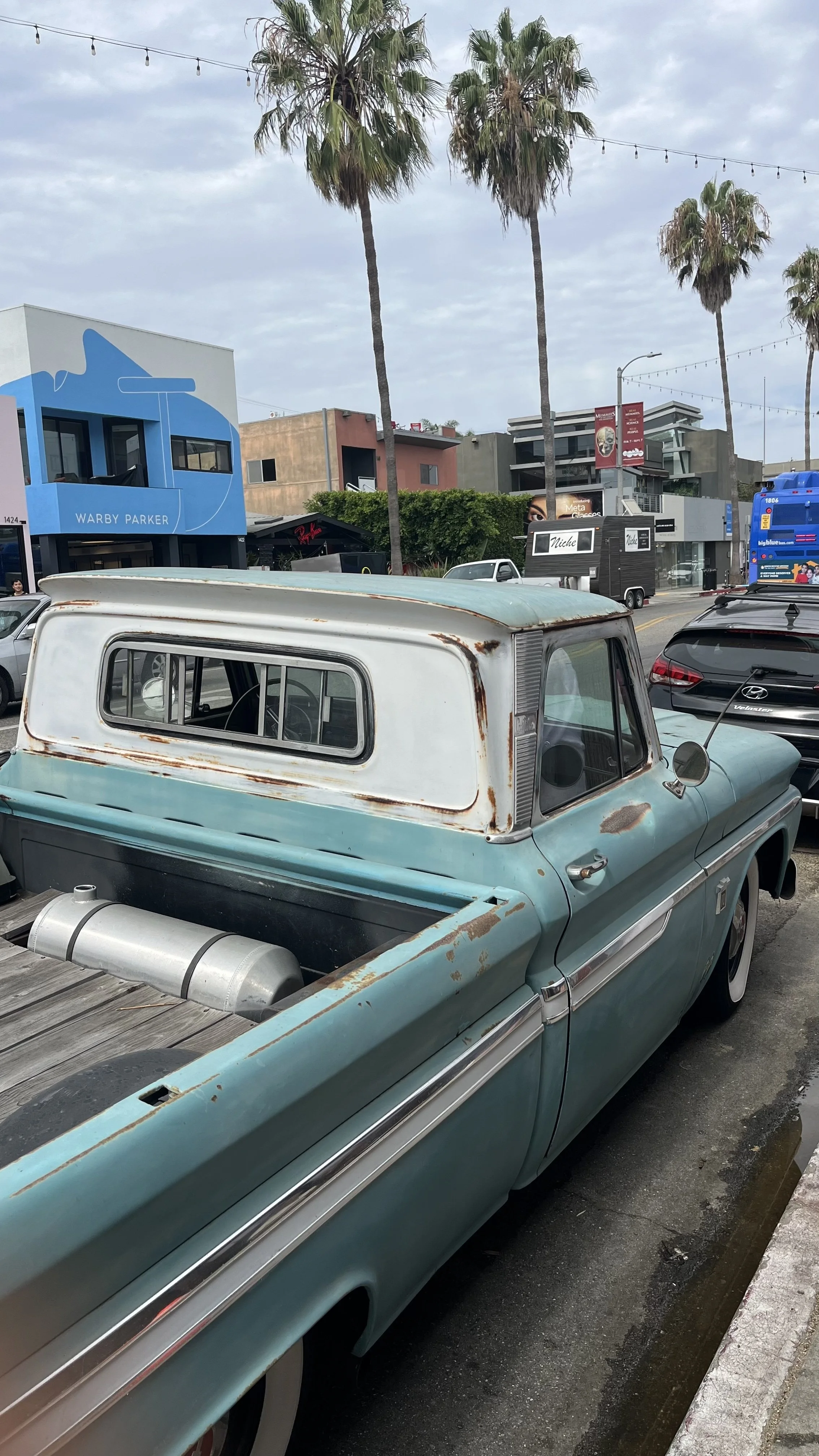 A vintage pickup truck with rust and chipped paint, parked in front of a shopping district with modern buildings and tall palm trees.