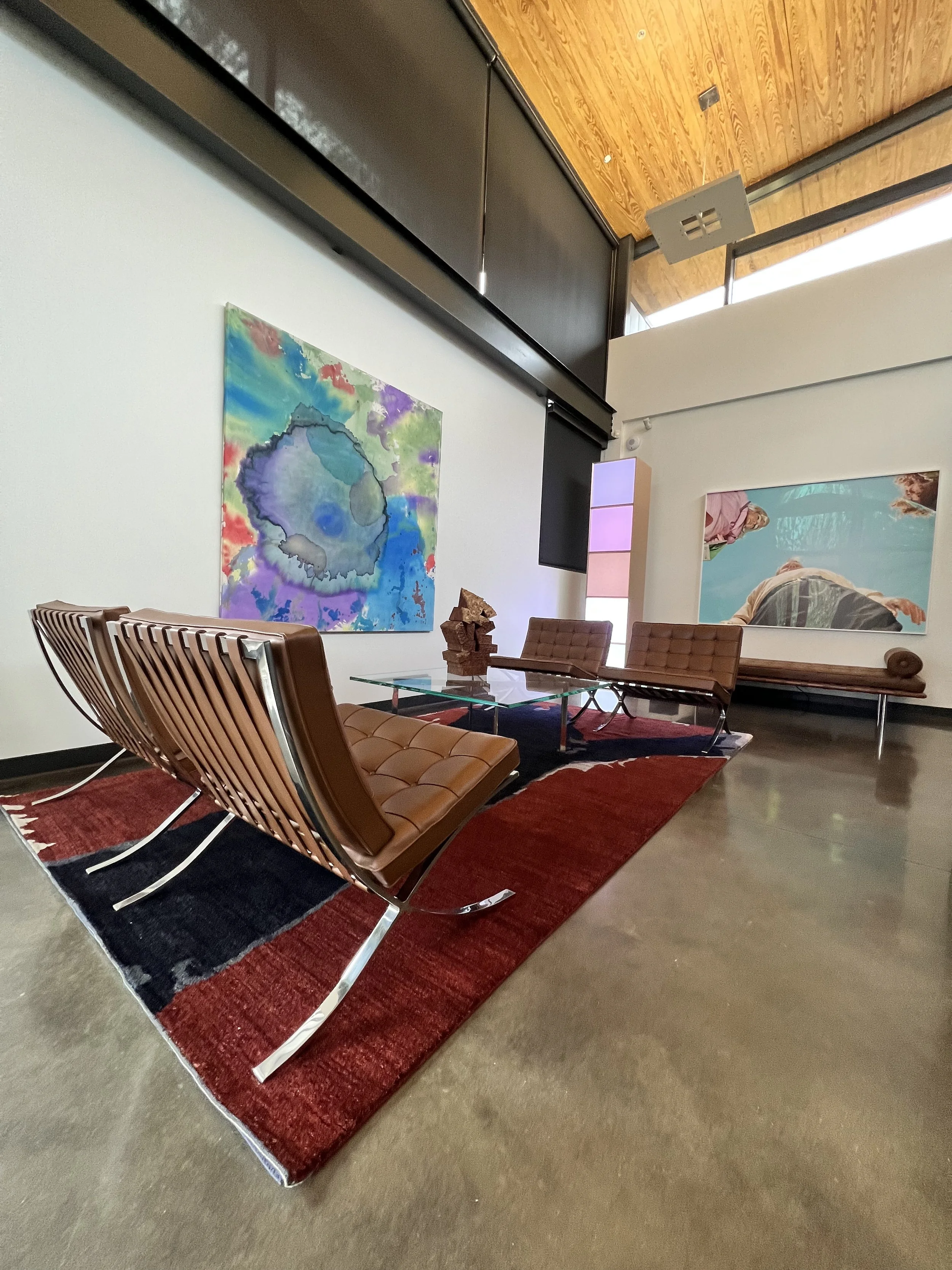 Interior of an art gallery or museum with abstract paintings on white walls, a glass coffee table with decorative objects, two wooden chairs with brown cushions, a red and black rug, and a wooden ceiling with skylights.