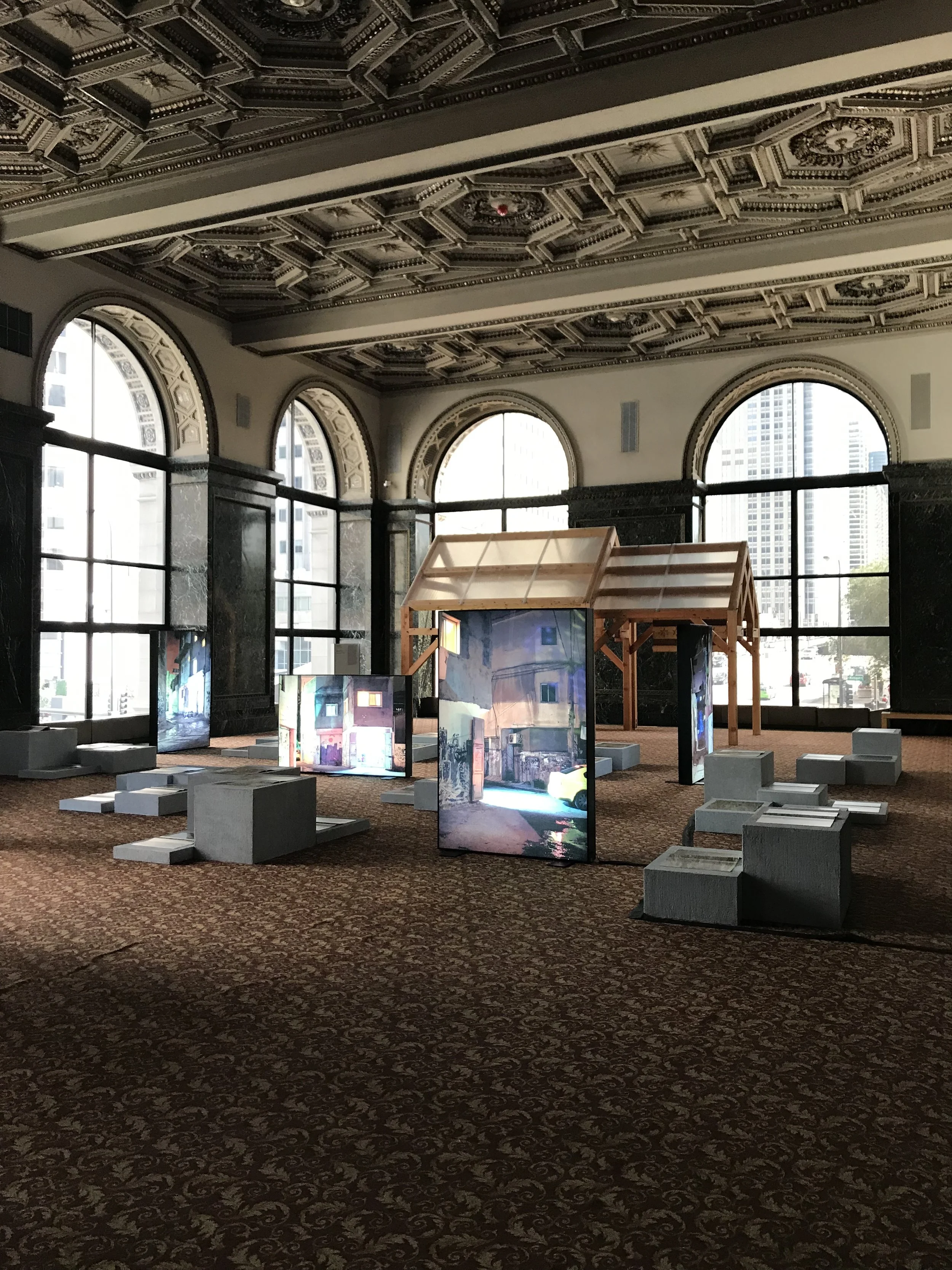 Exhibition space in a grand hall with ornate ceiling, large arched windows, and cityscape outside. Art displays include digital screens showing city scenes and a wooden structure.