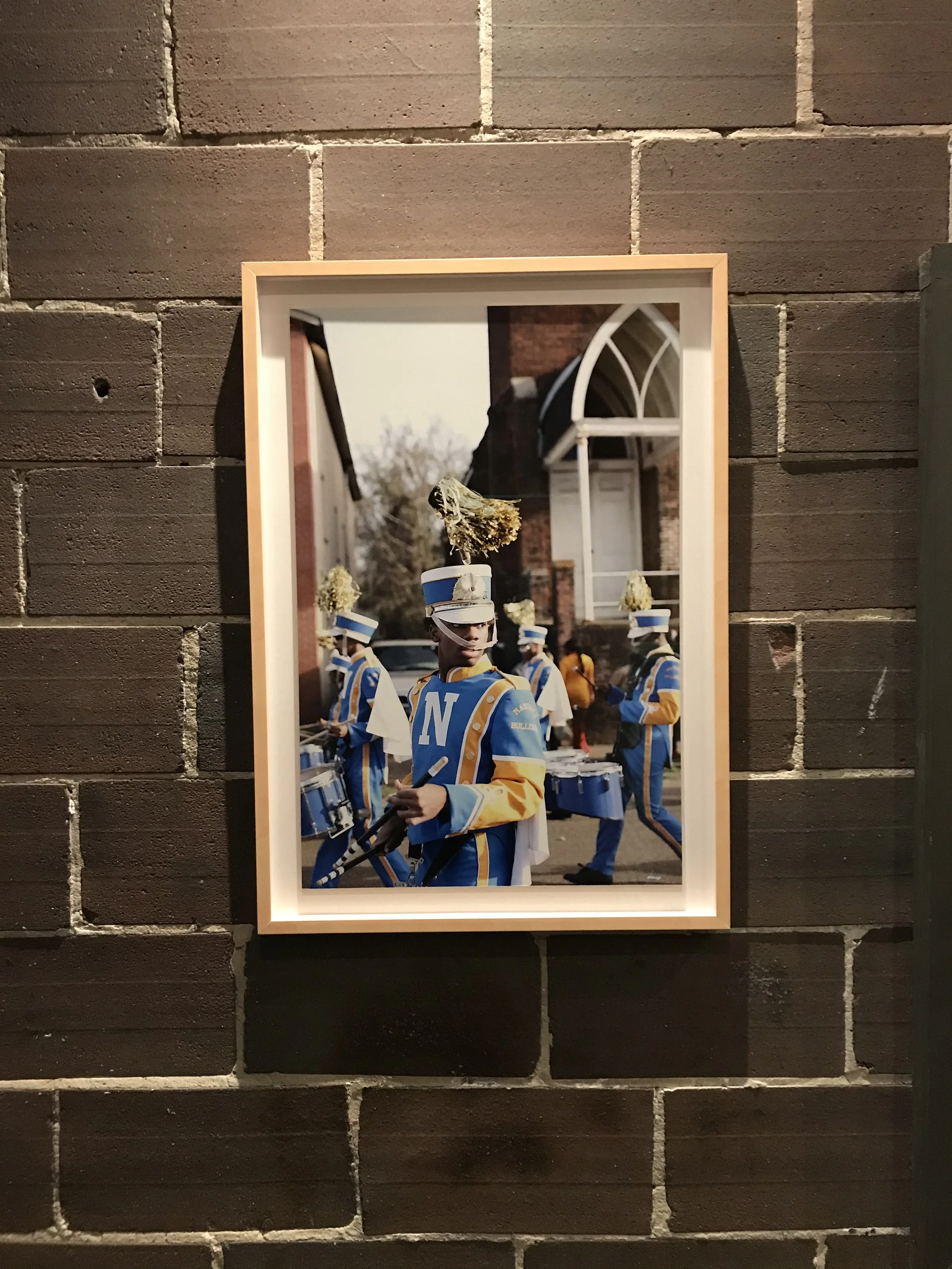 Photo of a marching band in blue and yellow uniforms playing drums, standing outdoors in front of a brick building.