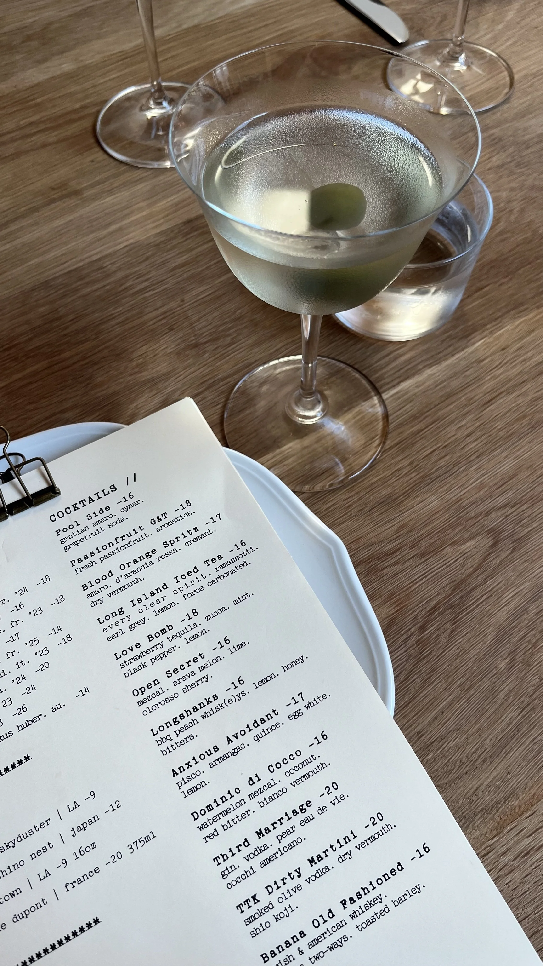 A menu on a white plate on a wooden table, with two glasses of water and a cocktail glass with a clear drink and green olives.