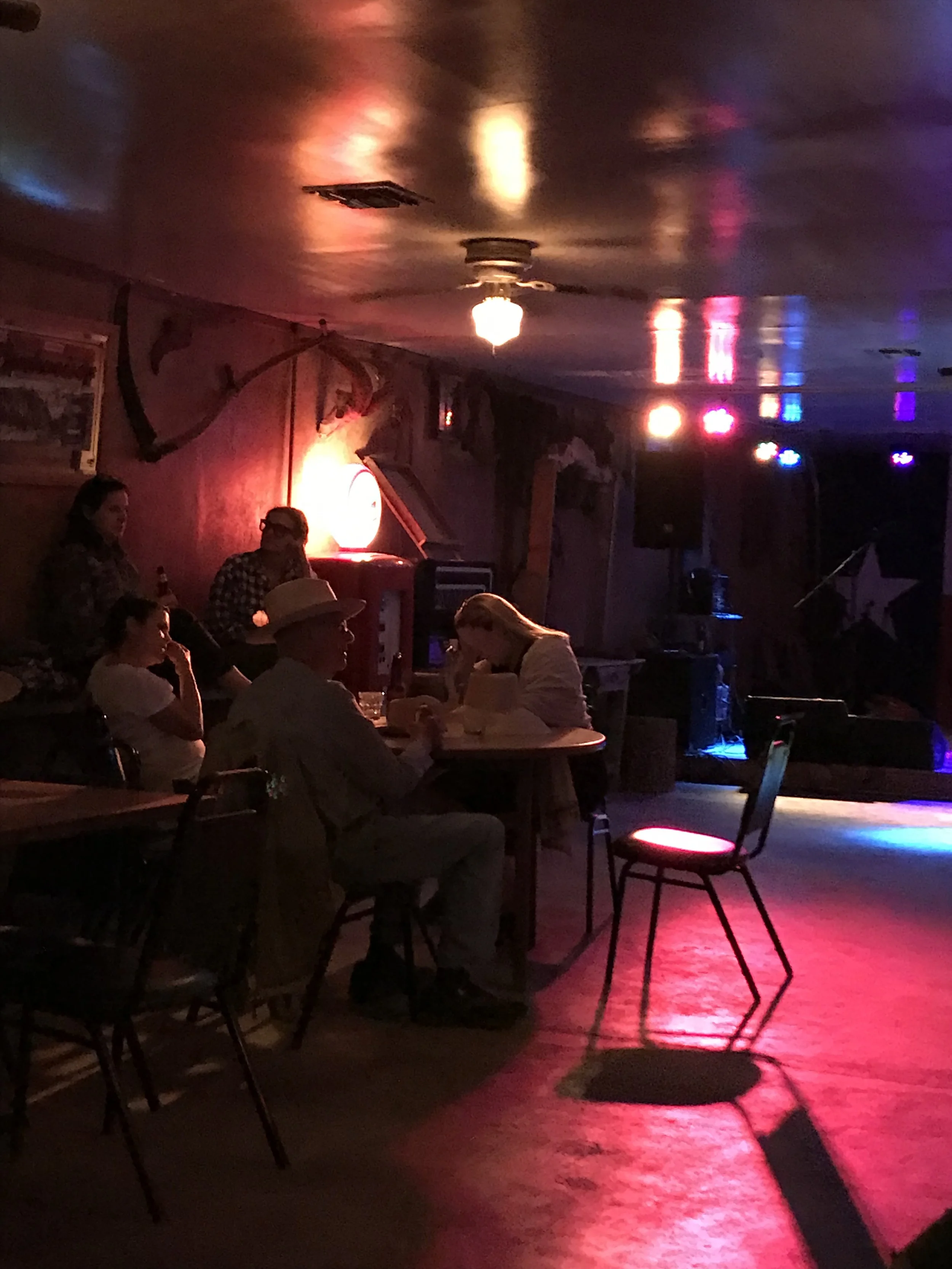 A dimly lit indoor bar or restaurant with a group of people sitting around tables, some wearing cowboy hats, and a ceiling fan with colorful flashing lights reflecting off the ceiling.