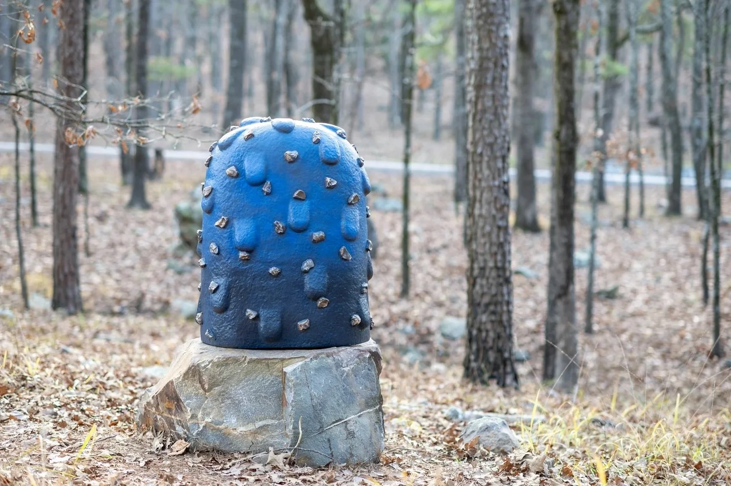 Blue, rounded sculpture with protruding metal studs, mounted on a large rock, in a wooded area with trees and fallen leaves.