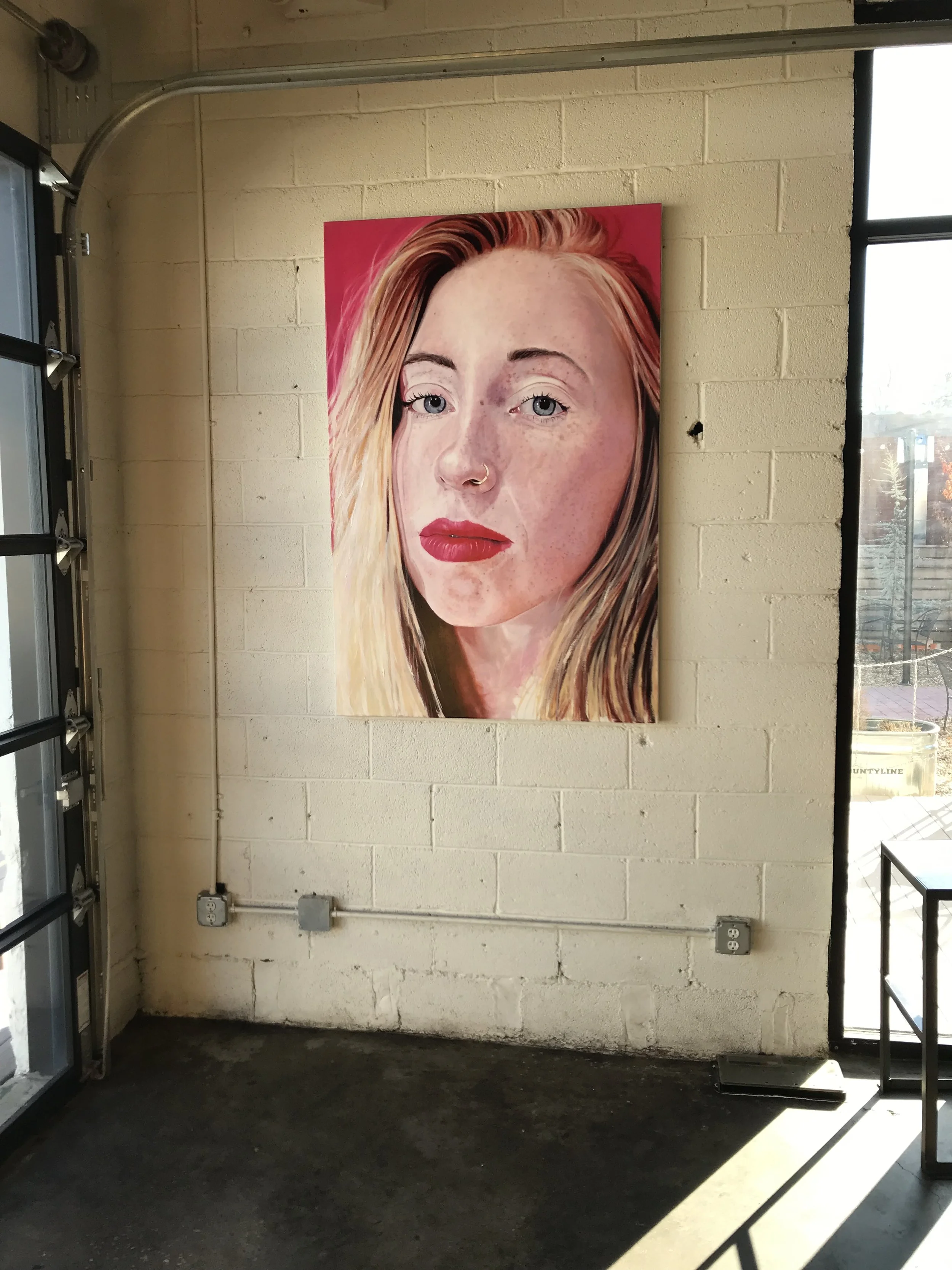 A large, colorful portrait of a woman with blonde hair and red lipstick is hanging on a brick wall in an industrial-style room with a concrete floor and natural light coming through the window.