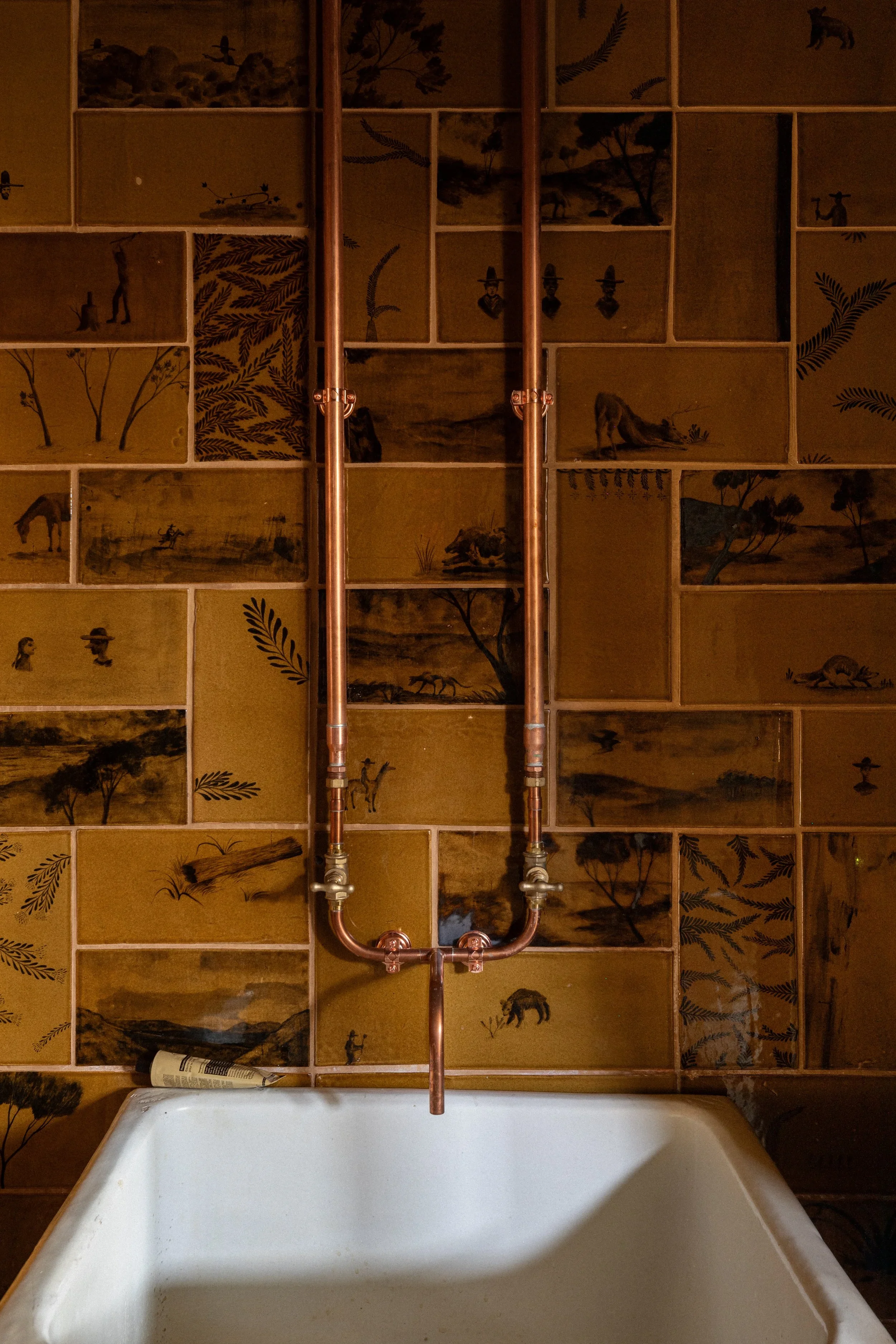 A bathroom with a vintage style copper pipe faucet above a white bathtub. The background features a decorative wall with a collage of vintage landscape and nature paintings or prints, some depicting trees, animals, and figures.