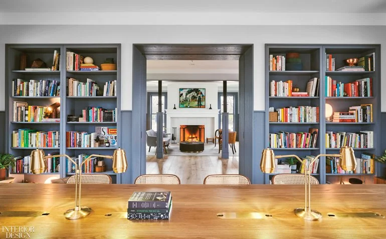 Library bookshelves, table, and a fireplace with artwork over it in the distance.
