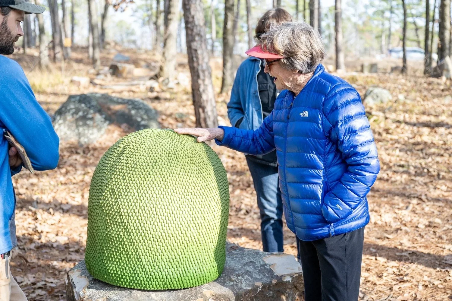 Older woman in a blue jacket and sunglasses touches a large, textured, yellow-green sculpture in a wooded outdoor area while two men watch.