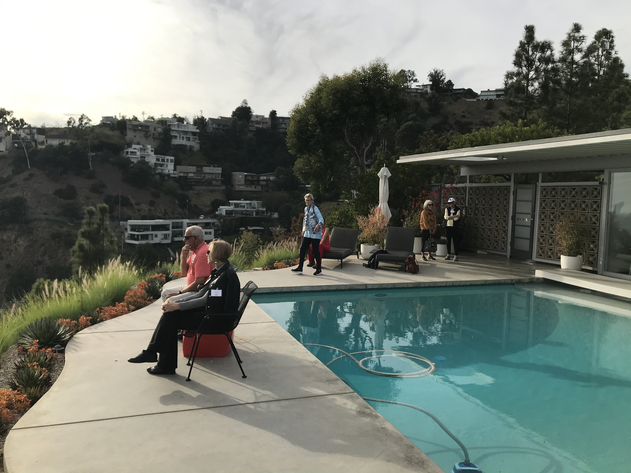 People relaxing by a swimming pool with a view of hillside houses and trees in the background.
