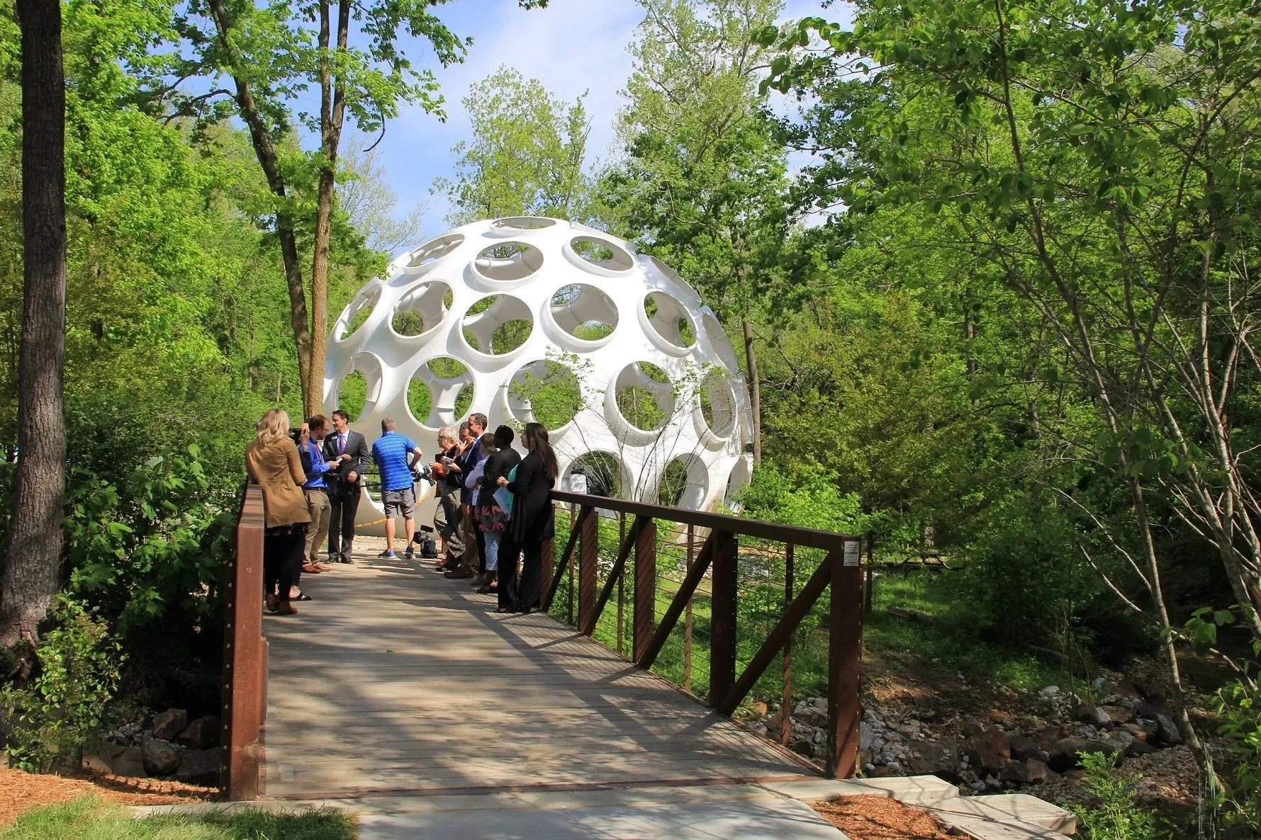 People gathered on a wooden walkway in a lush green forest, with a large white geodesic sphere structure in the background.