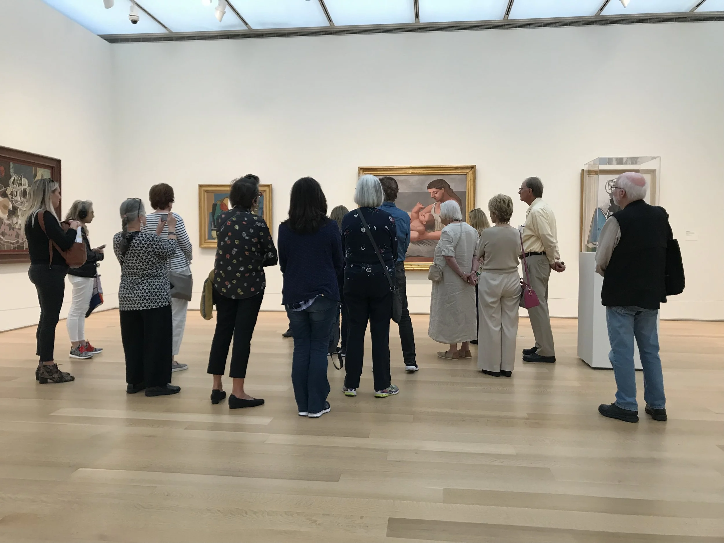 A group of people gathered in an art gallery, looking at paintings on the wall. The gallery has white walls and wooden floors.