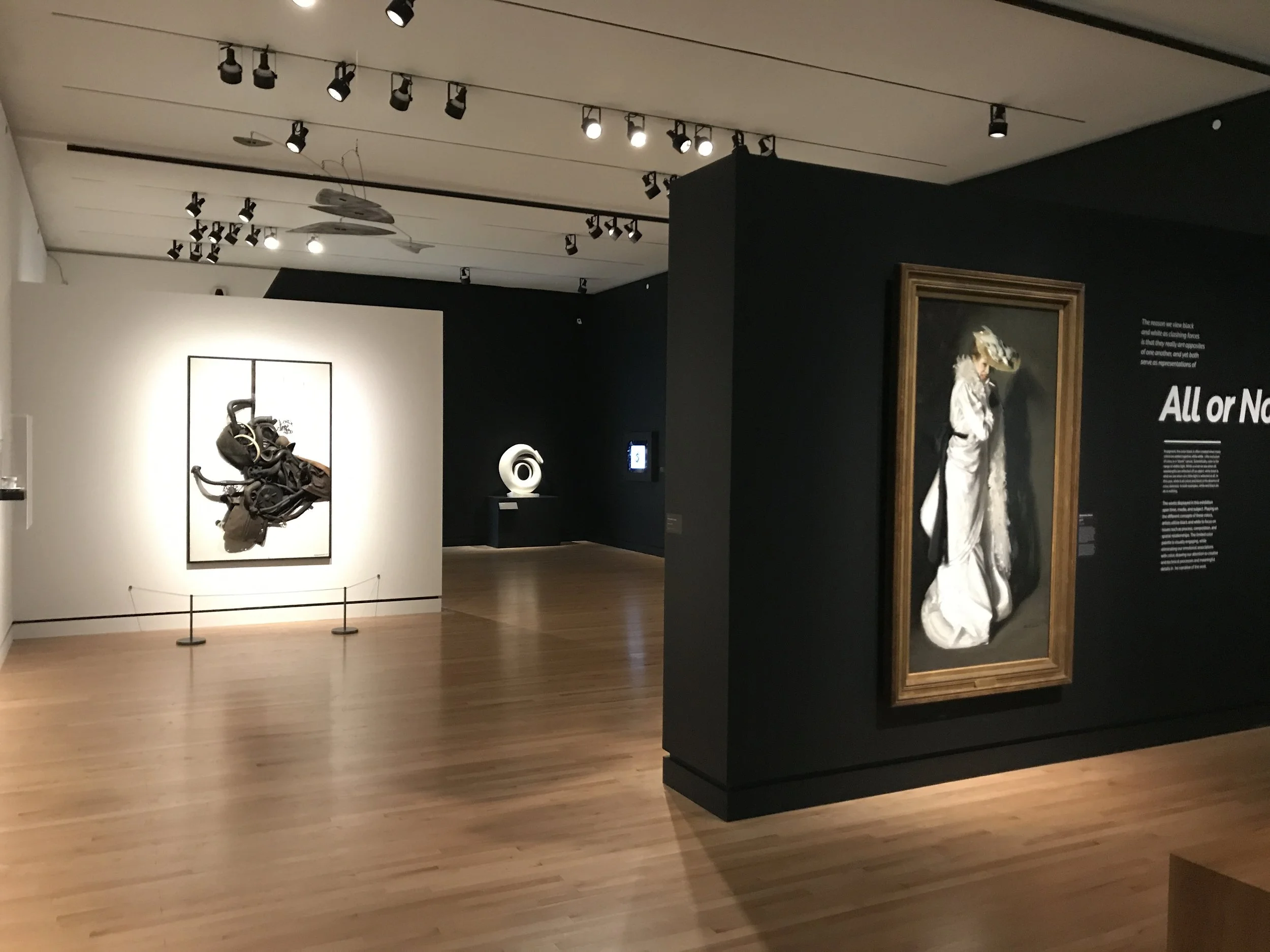 View inside a museum or gallery with artworks and sculptures, including a framed sculpture of a figure in a white outfit with a large hat, displayed on a black partition. The room has wooden flooring, track lighting on the ceiling, and white and dark-colored walls with descriptive text.