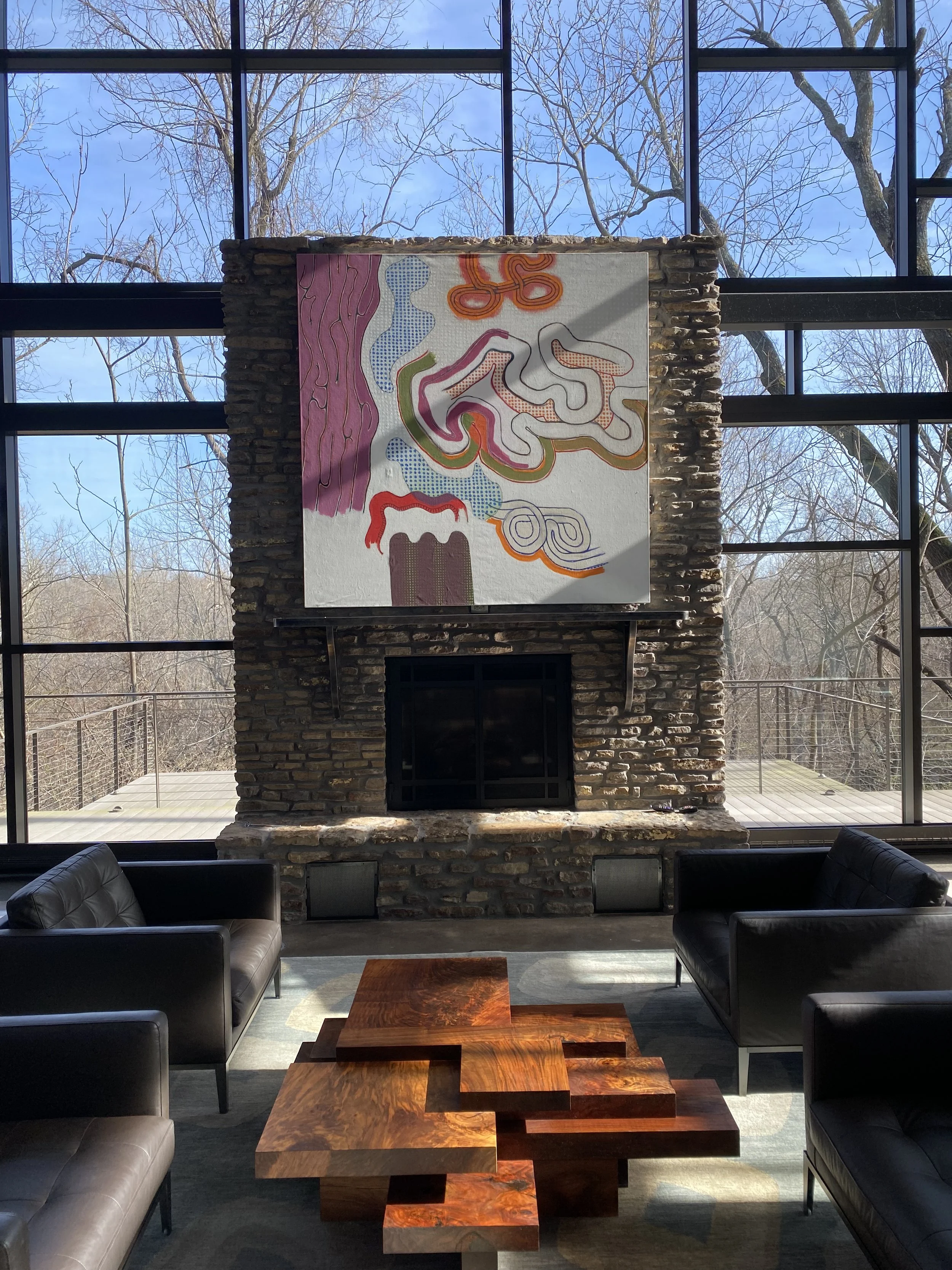 Interior view of a living room with a large abstract art piece hanging above a stone fireplace. The room has large floor-to-ceiling windows showing leafless trees and blue sky outside. There are black armchairs and a modern wooden coffee table in the room.