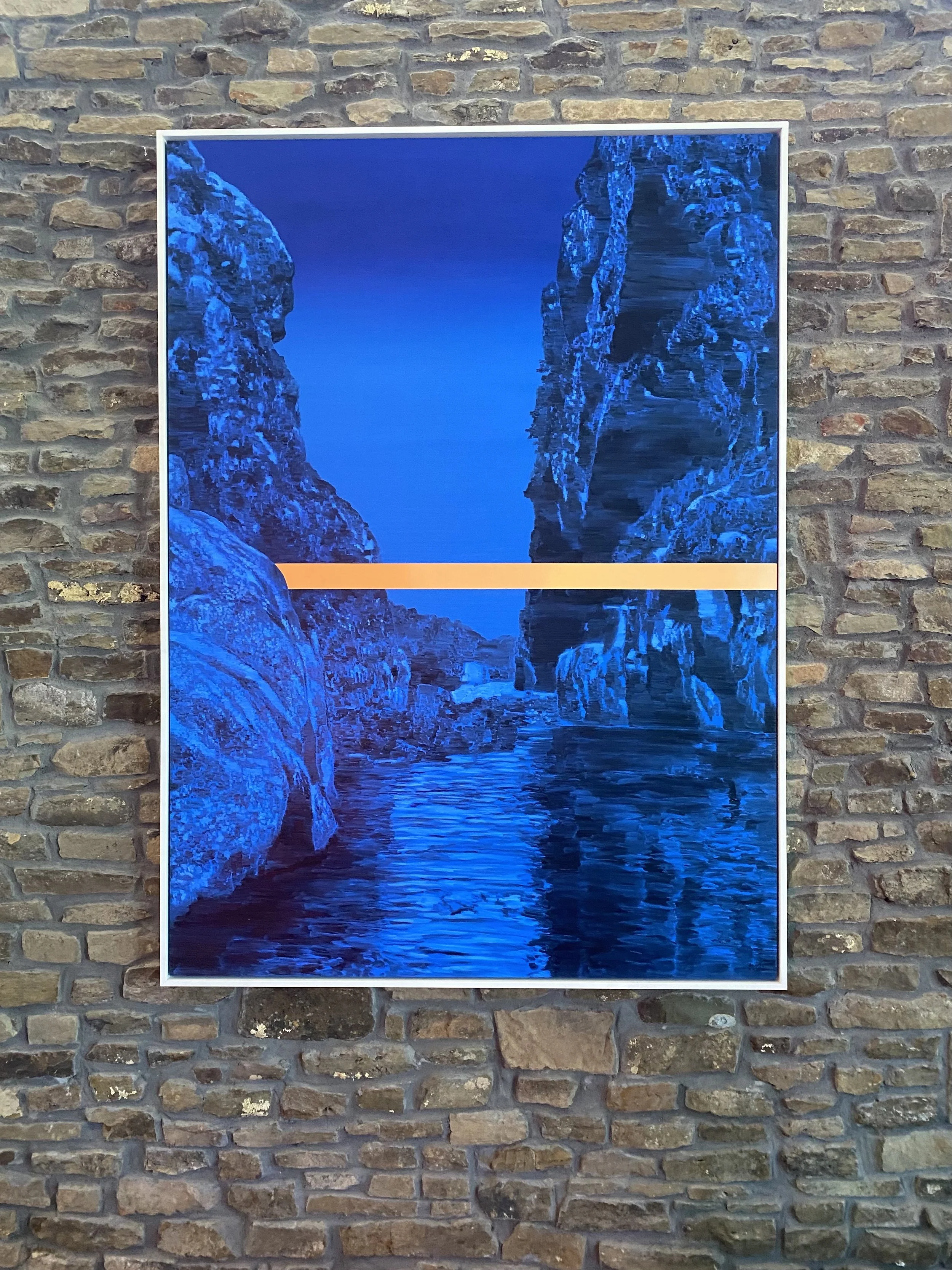 A framed photograph of a deep blue canyon with rocky formations and a body of water, mounted on a brick wall.