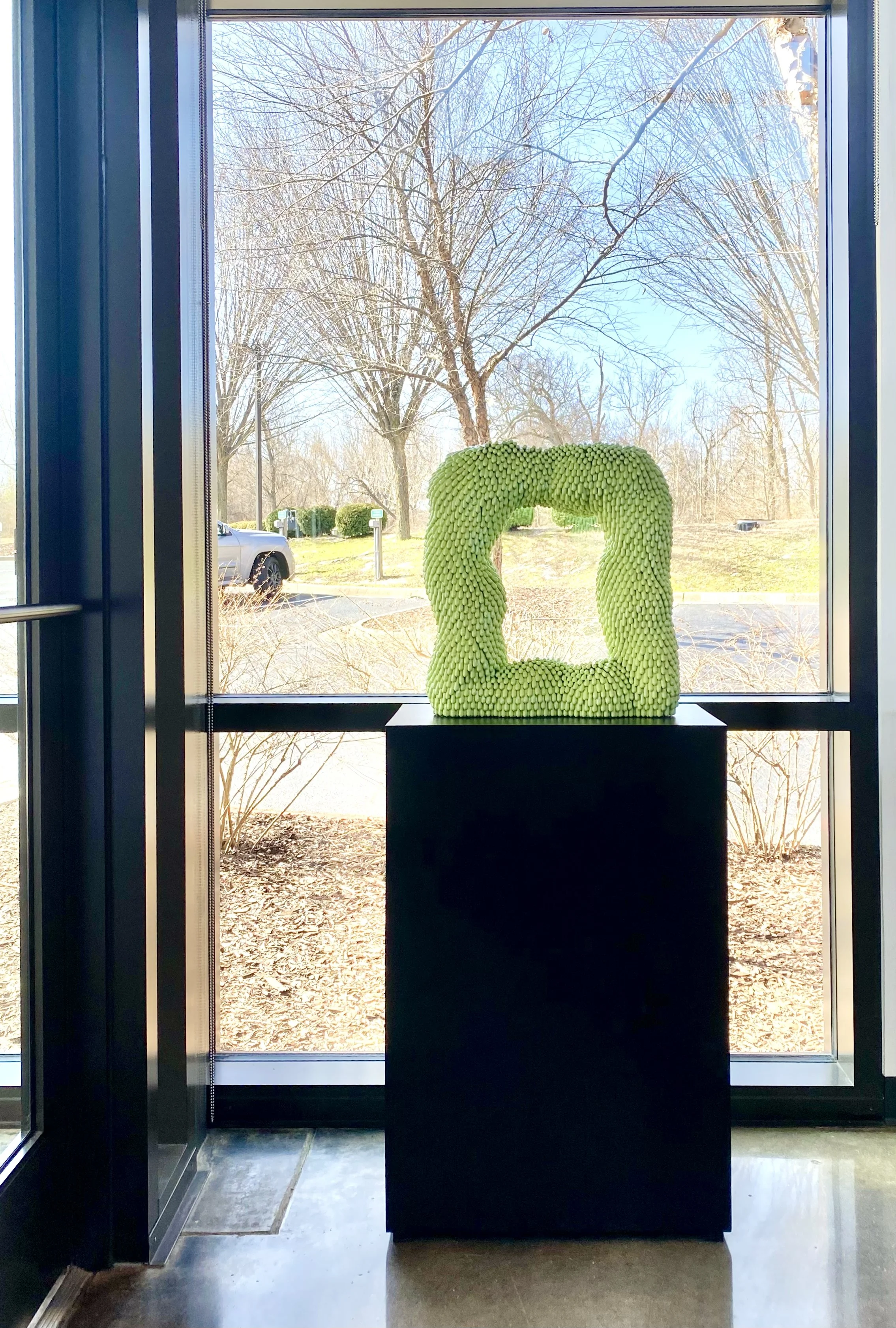 Green sculpture made of numerous small, elongated shapes, displayed on a black pedestal against a window with an outdoor scene of leafless trees and a parking lot.