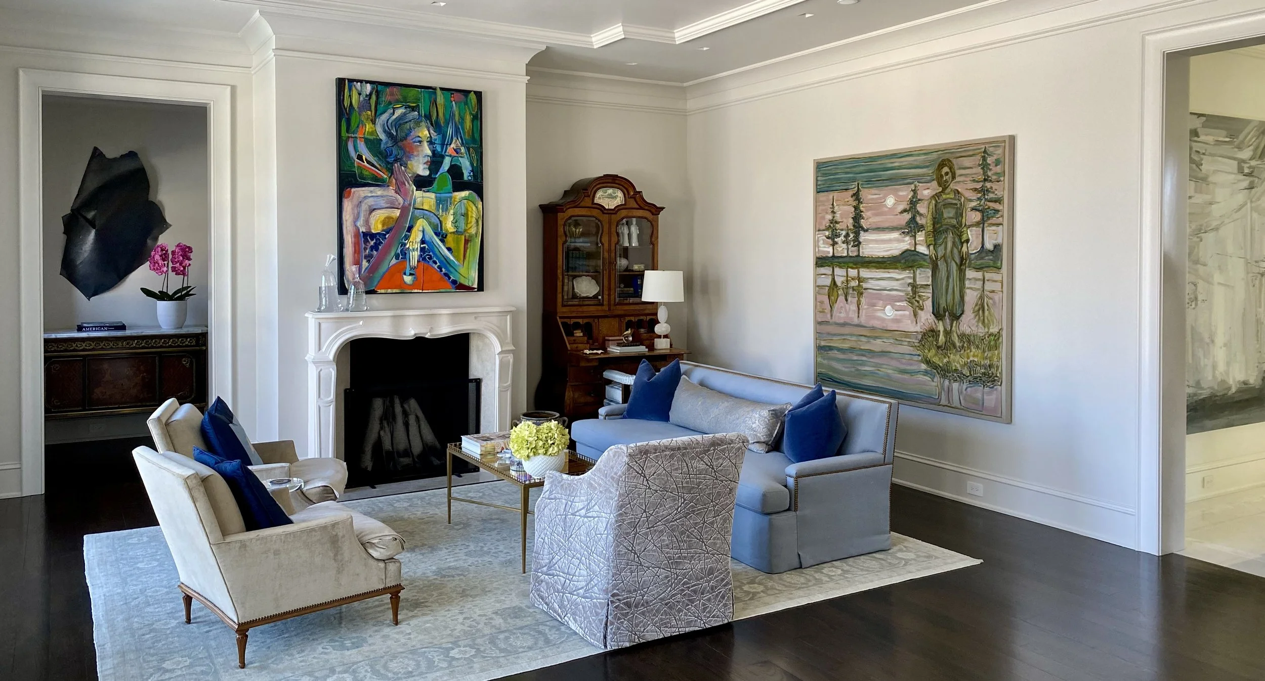 Living room with white walls, dark hardwood floors, a fireplace, and colorful artwork including a large portrait of a woman and a landscape scene. Furniture includes blue and beige sofas, a dining chair, a matching armchair, pillows, a coffee table with a flower arrangement, and various decorative items and lamps.