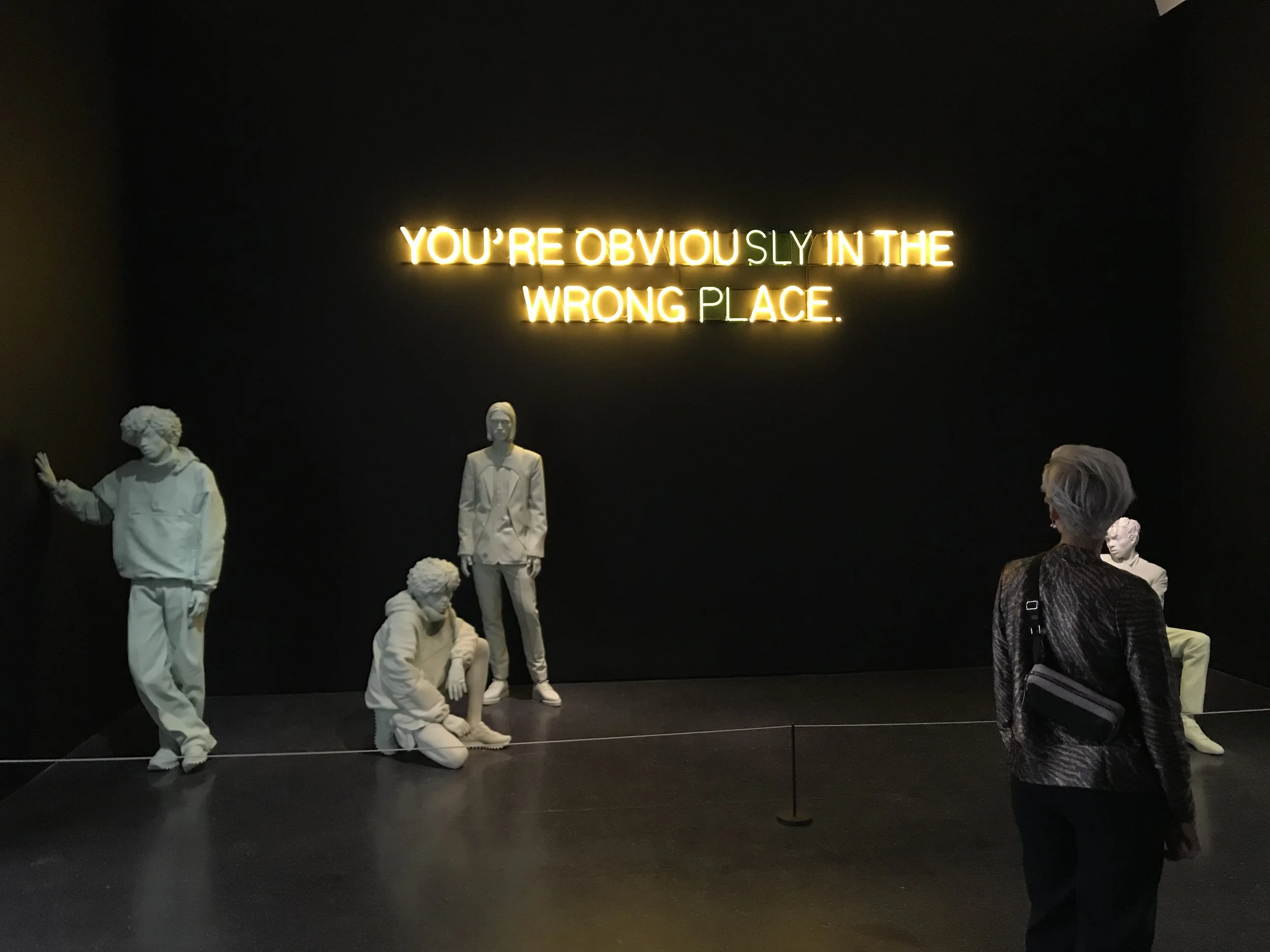 Neon sign in yellow and white reads, "YOU'RE OBVIOUSLY IN THE WRONG PLACE" on a black wall. Several white mannequins in casual clothing are positioned in front of the wall, with some seated and one standing, and a woman with glasses and a patterned jacket stands near the mannequins looking at them.