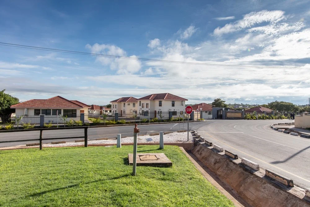 Residential neighborhood with houses featuring red-tile roofs, a stop sign at an intersection, a grassy area in the foreground, and a partly cloudy sky.