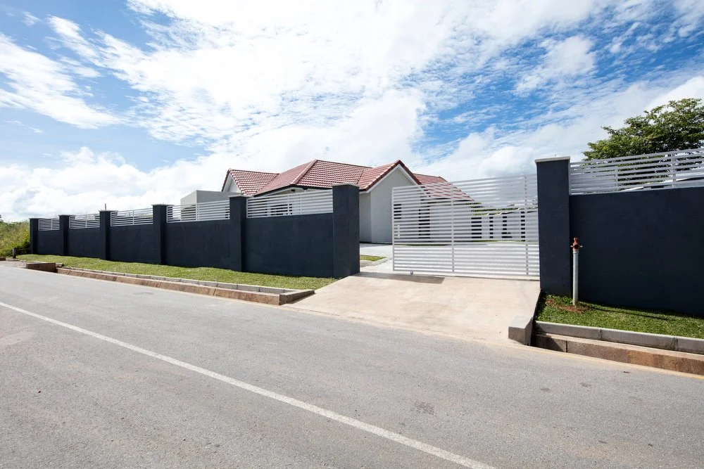 Modern house in Roma Park with a red roof behind a tall dark gray and white fence, front sidewalk and street, partly cloudy sky.