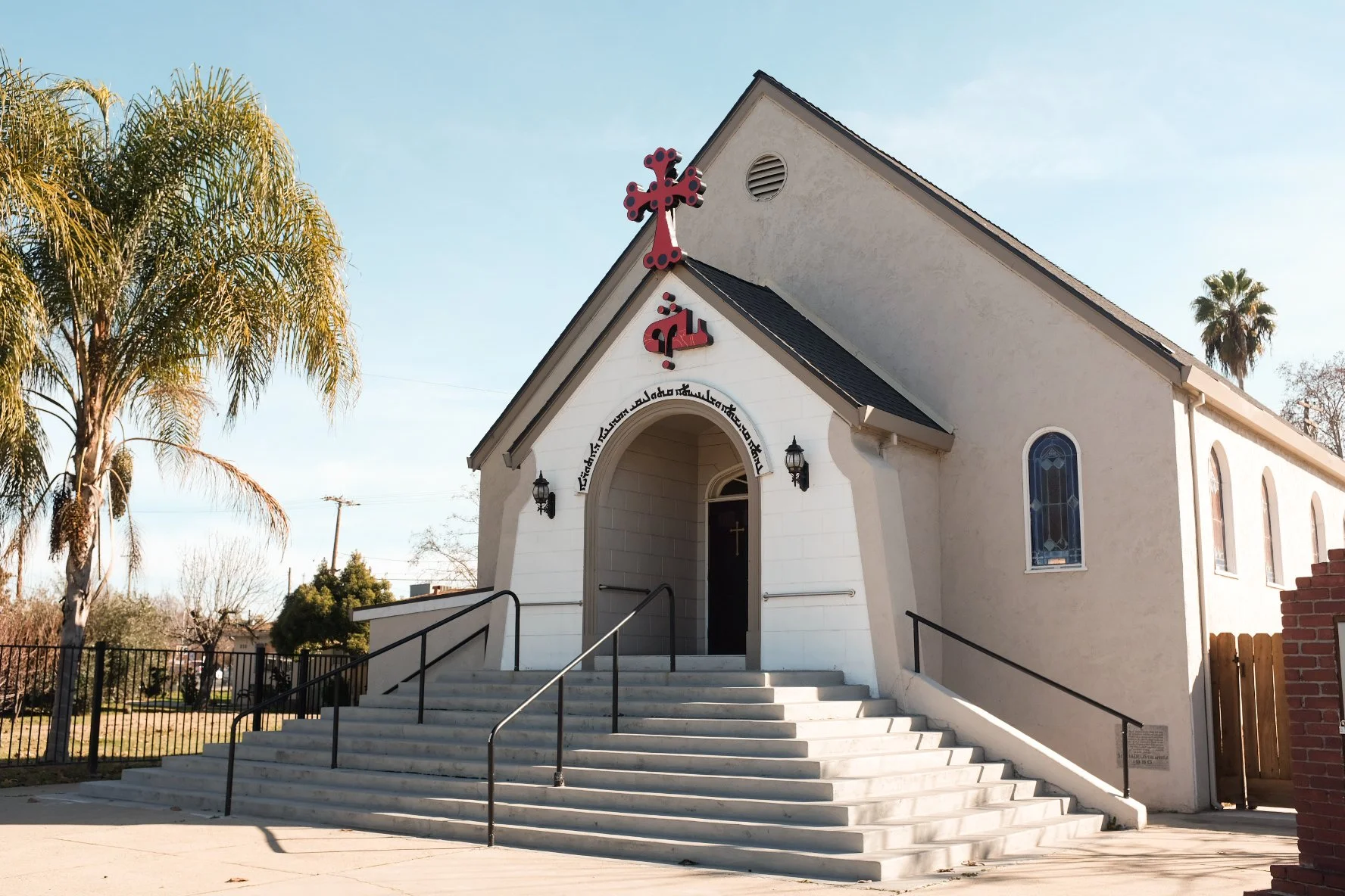 Front view of a small white church with steps leading up to the entrance, a red cross on top, and palm trees in the background.