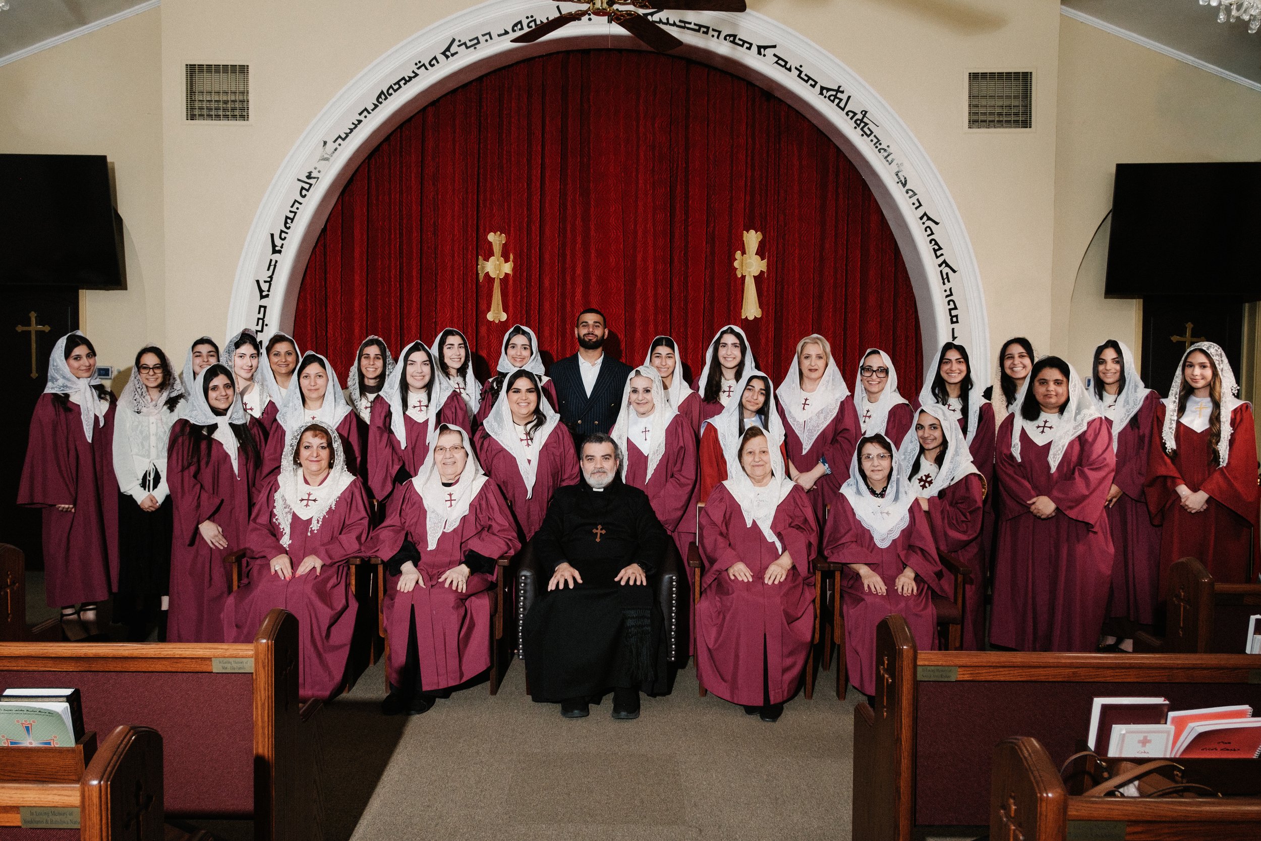 Group photo of individuals dressed in maroon robes and white head coverings, seated and standing in a church with red curtains and gold cross symbols in the background.