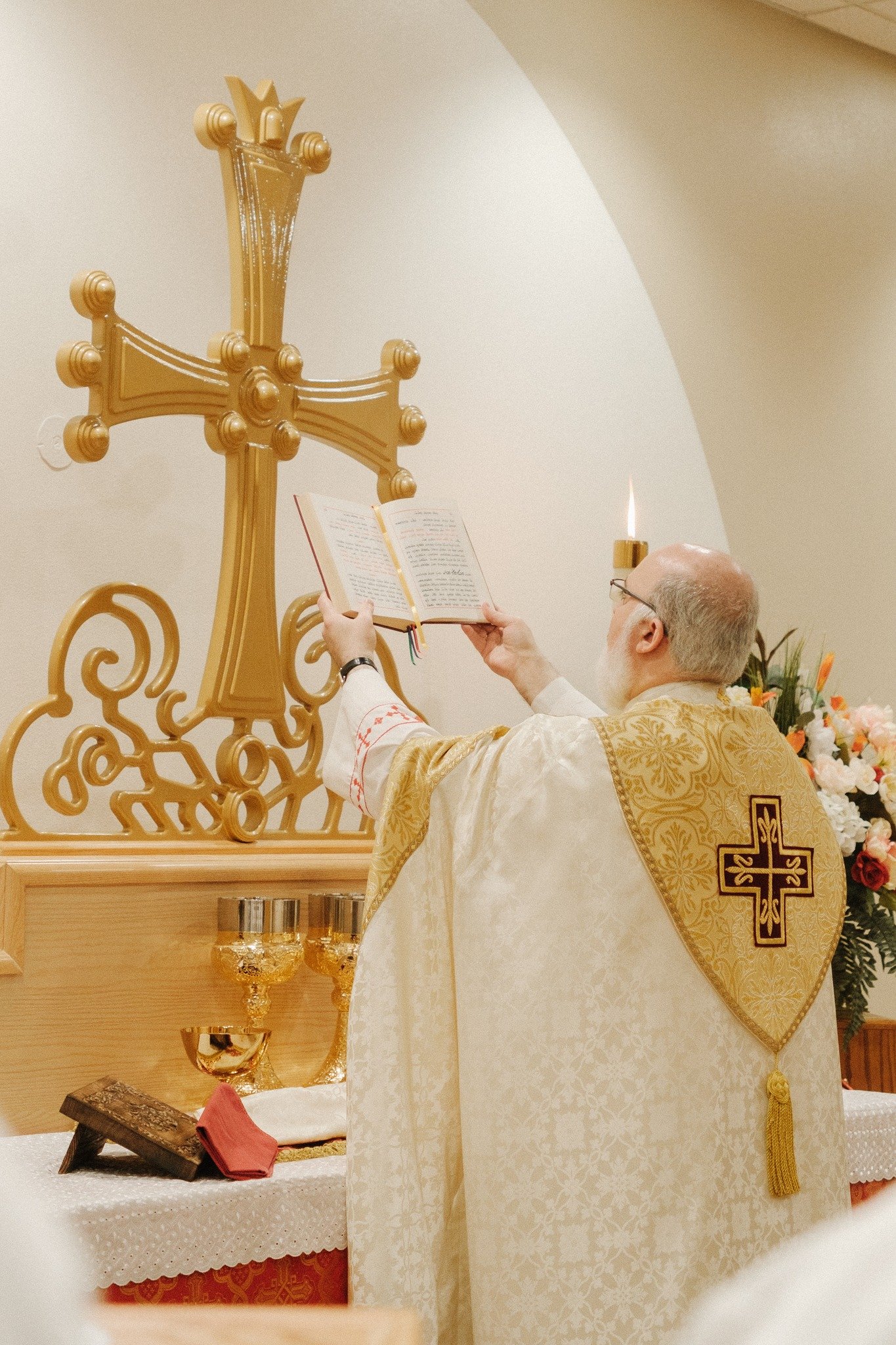 A priest in ornate vestments standing at an altar, holding an open religious book, during a Christian ceremony, with a large gold cross and floral arrangements in the background.