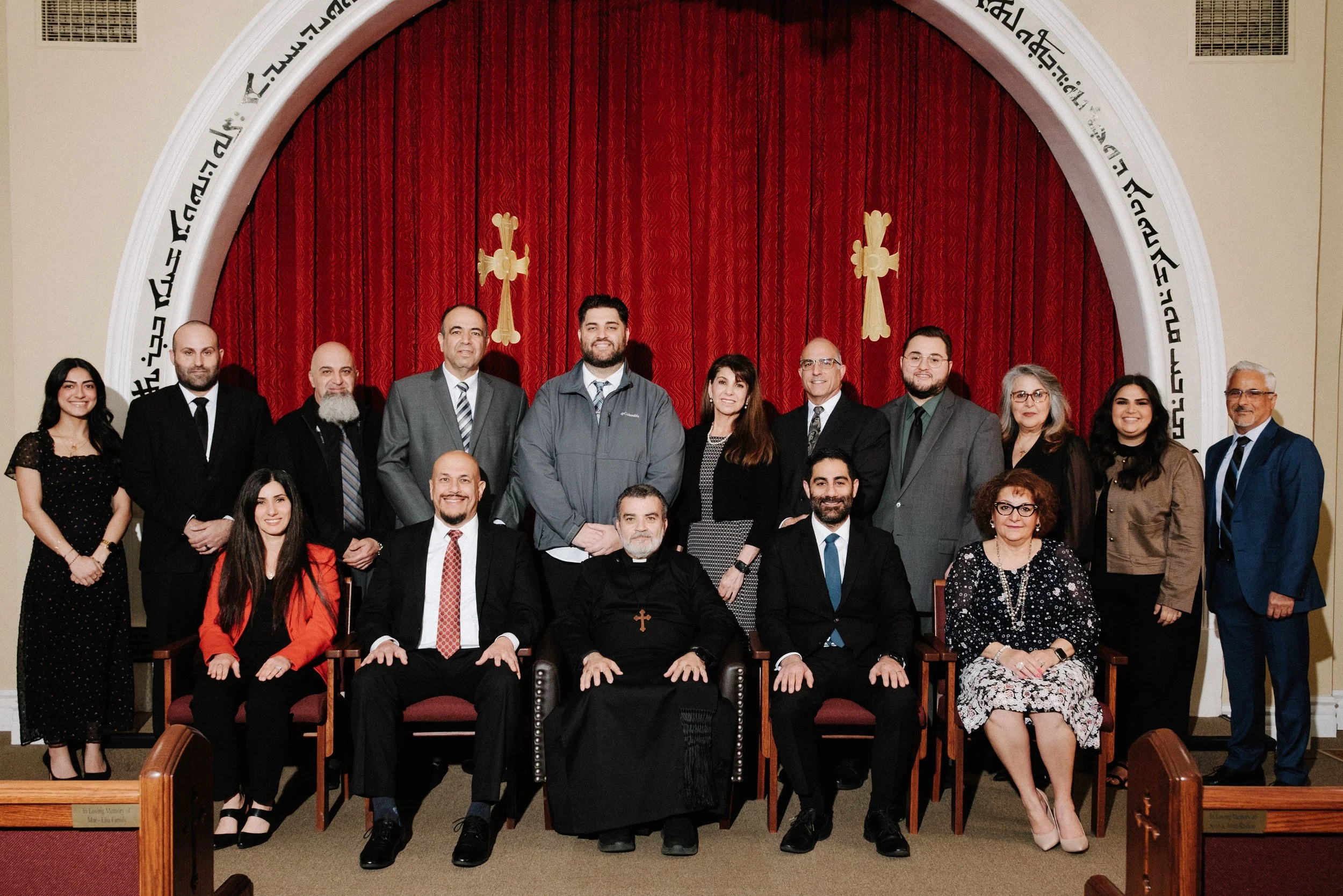 Group photo of 16 people, including clergy and laity, in a church with a red curtain and gold crosses in the background, posing for a picture.