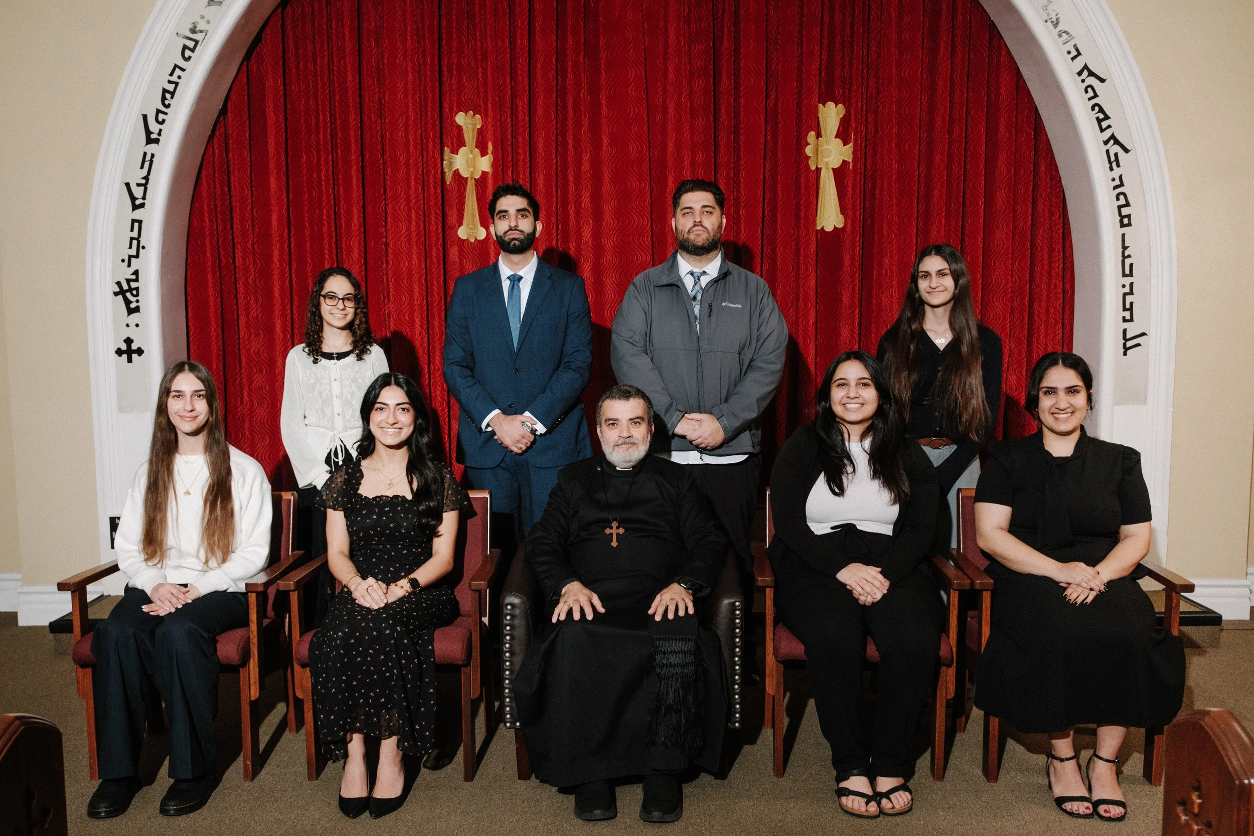 Group photo of ten people, including a priest in black robes with a cross necklace, posing in church with red curtains and gold crosses in the background. Four women are seated in front, and three men and three women are standing behind them.