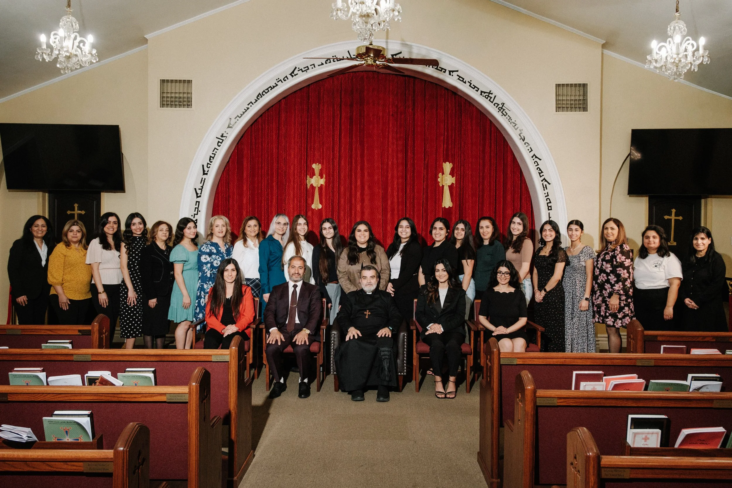 Group of people in a church sanctuary posing for a photo. The group includes men and women, some seated and some standing in front of a red curtain with religious symbols. The background features chandeliers, a pulpit, and cross symbols.