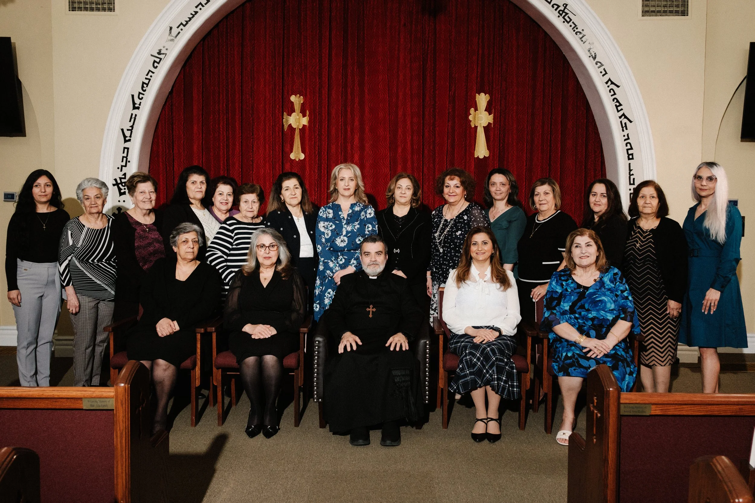 Group of women and one man, dressed in formal attire, seated and standing in front of a red curtain with religious symbols, inside a church or religious setting.
