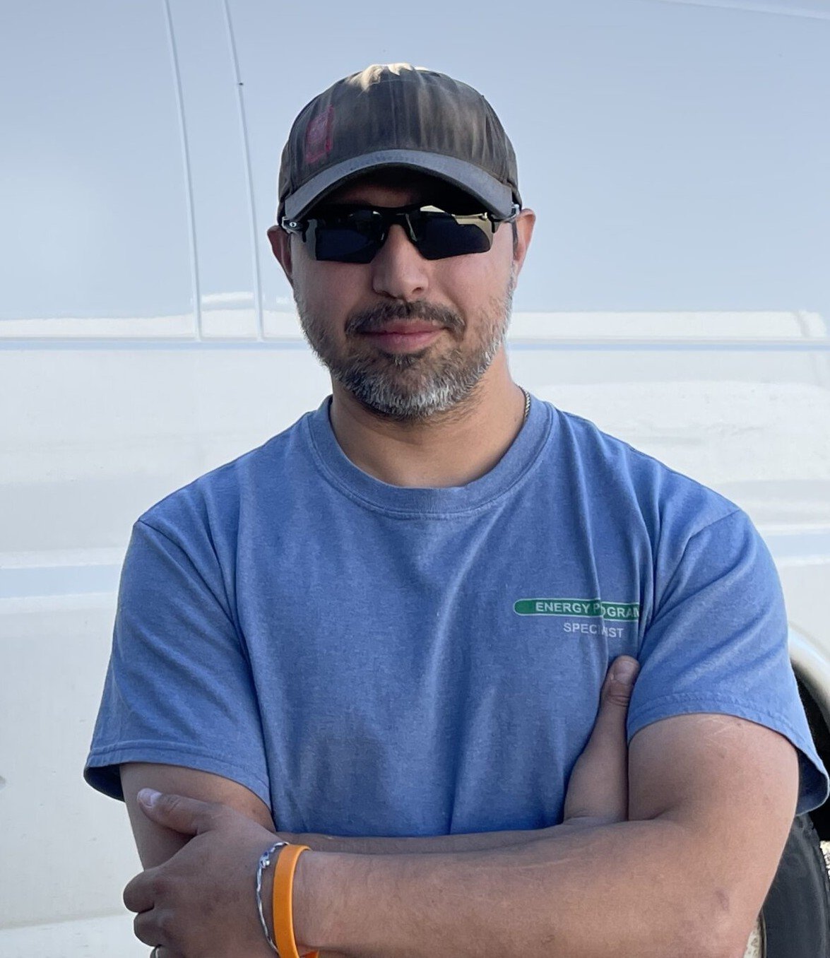 A man wearing sunglasses, a baseball cap, a gray t-shirt with a green logo on the chest, and standing with his arms crossed in front of a white vehicle.