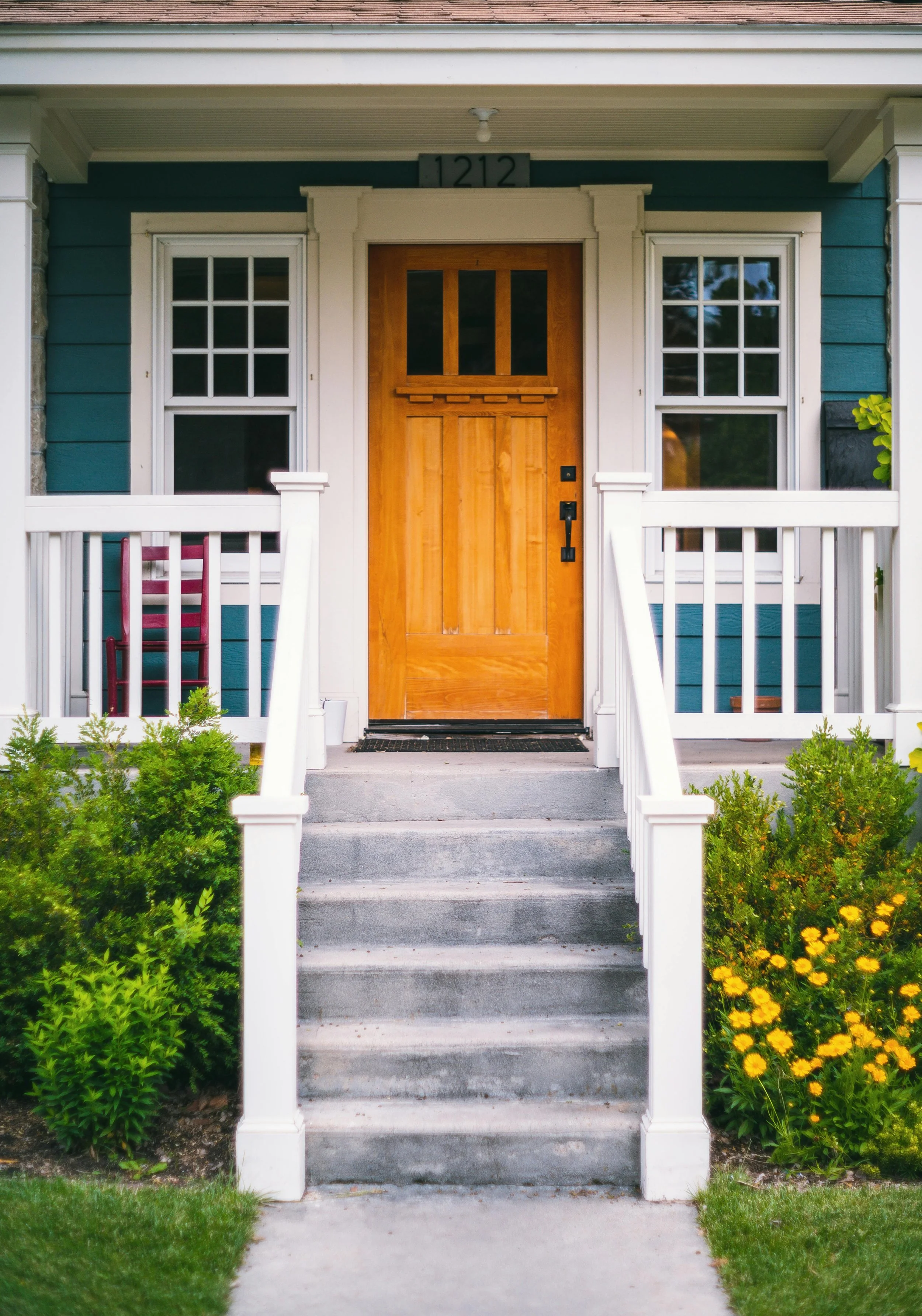 Front porch of a house with brick steps, white railing, a wooden front door with glass panes, two large windows, green bushes with yellow flowers, and house number 1212 above the door.