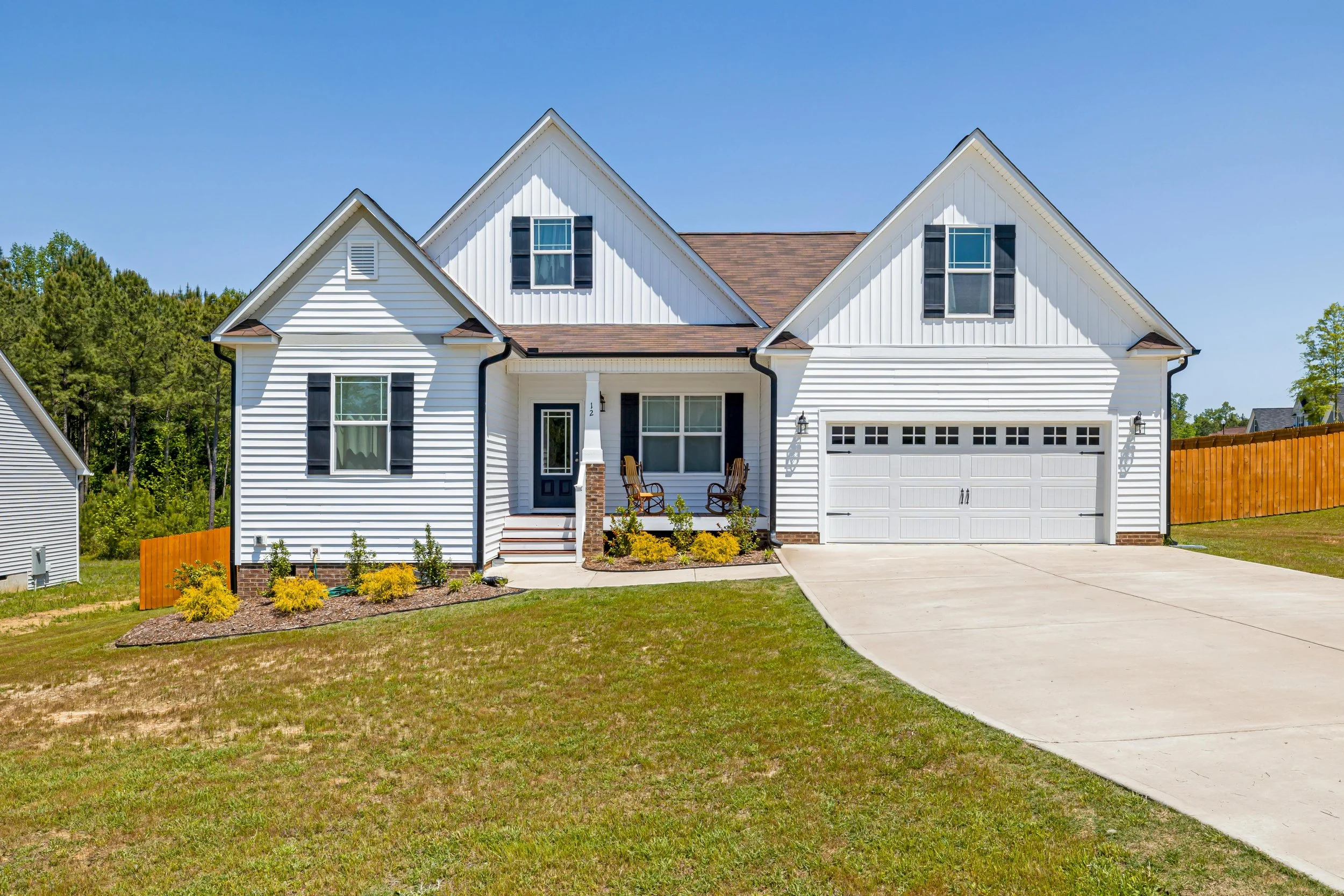 White two-story house with black shutters, attached garage, front porch with chairs, landscaped yard, blue sky, and neighboring houses in the background.