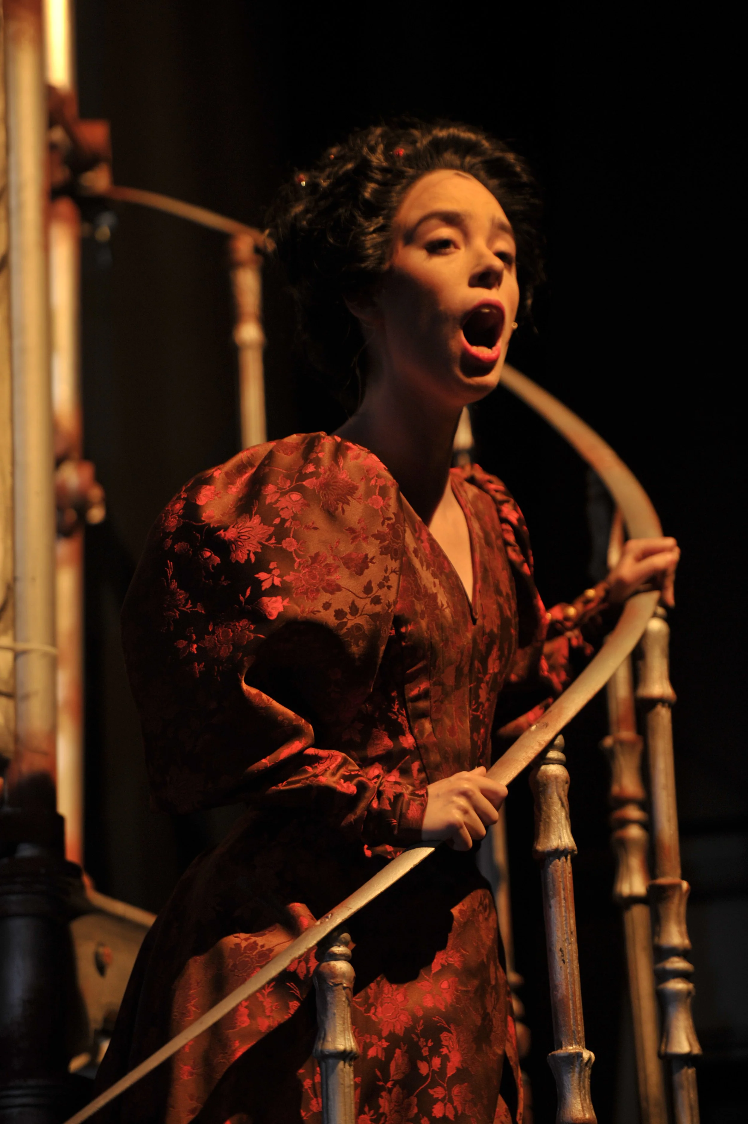 A woman in a red, floral-patterned dress is on a staircase, with her mouth open, possibly singing or yelling, with a dark background.