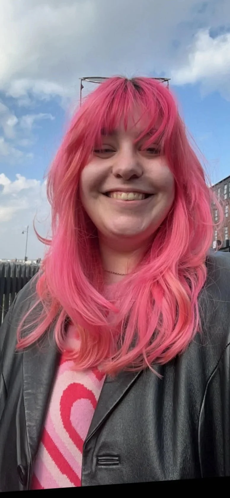 Smiling woman with pink hair, wearing glasses on her head and a black leather jacket, outdoors on a cloudy day.