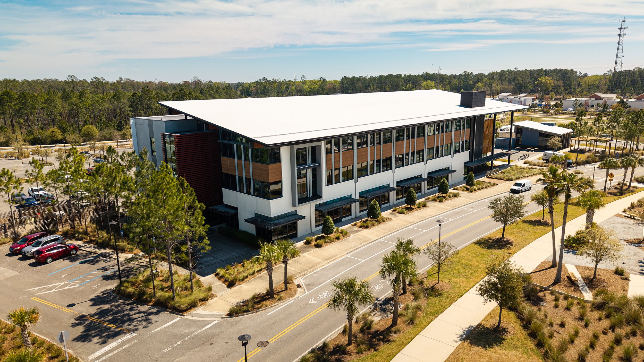 A modern, multi-story office building surrounded by a landscaped area with palm trees and a parking lot, seen from above on a sunny day.